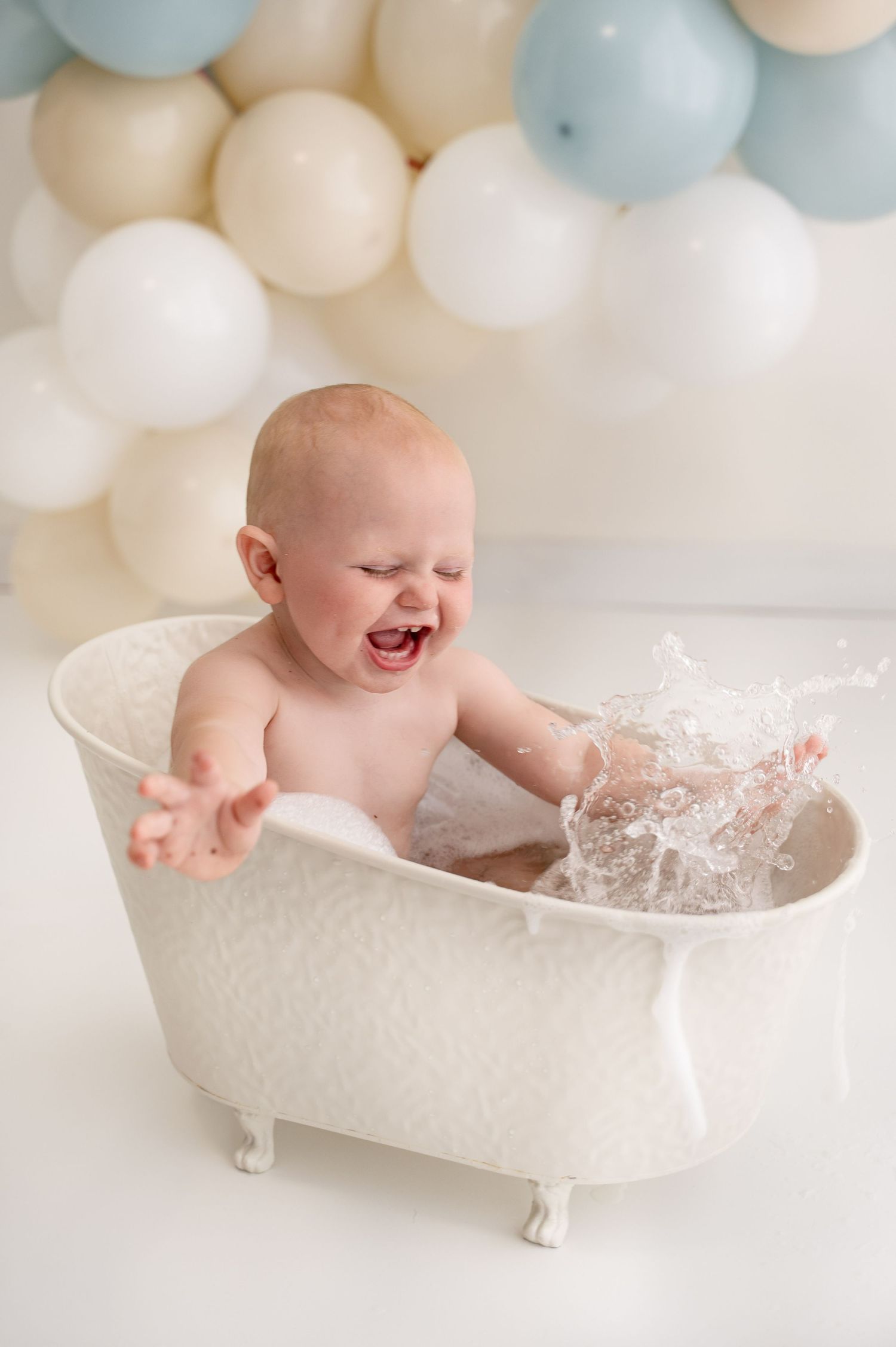 Baby splashing and playing in vintage white bathtub with balloon backdrop.