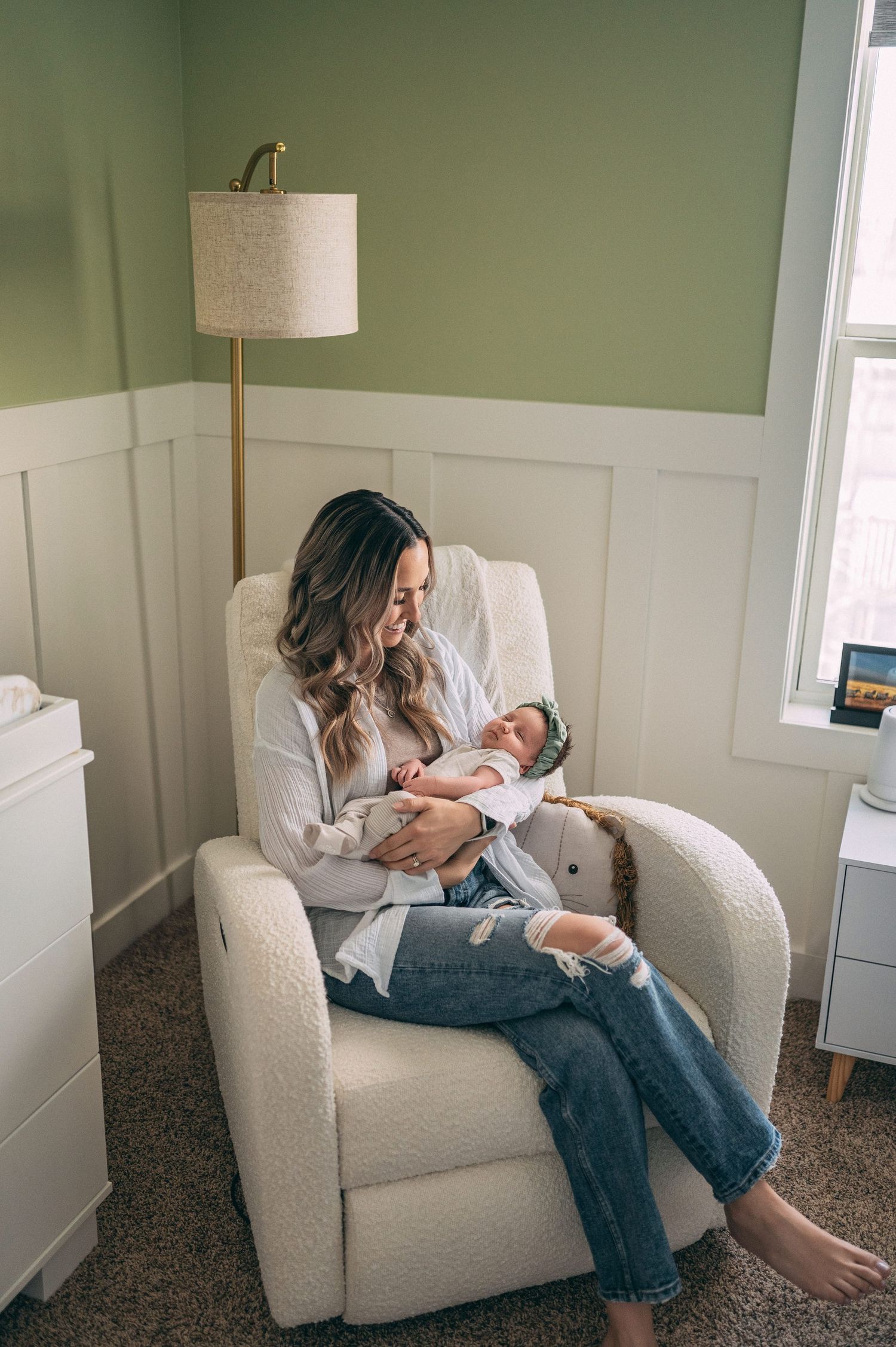 A parent sits in a cream-colored armchair in a nursery with sage green walls, tending to a newborn in a white room.
