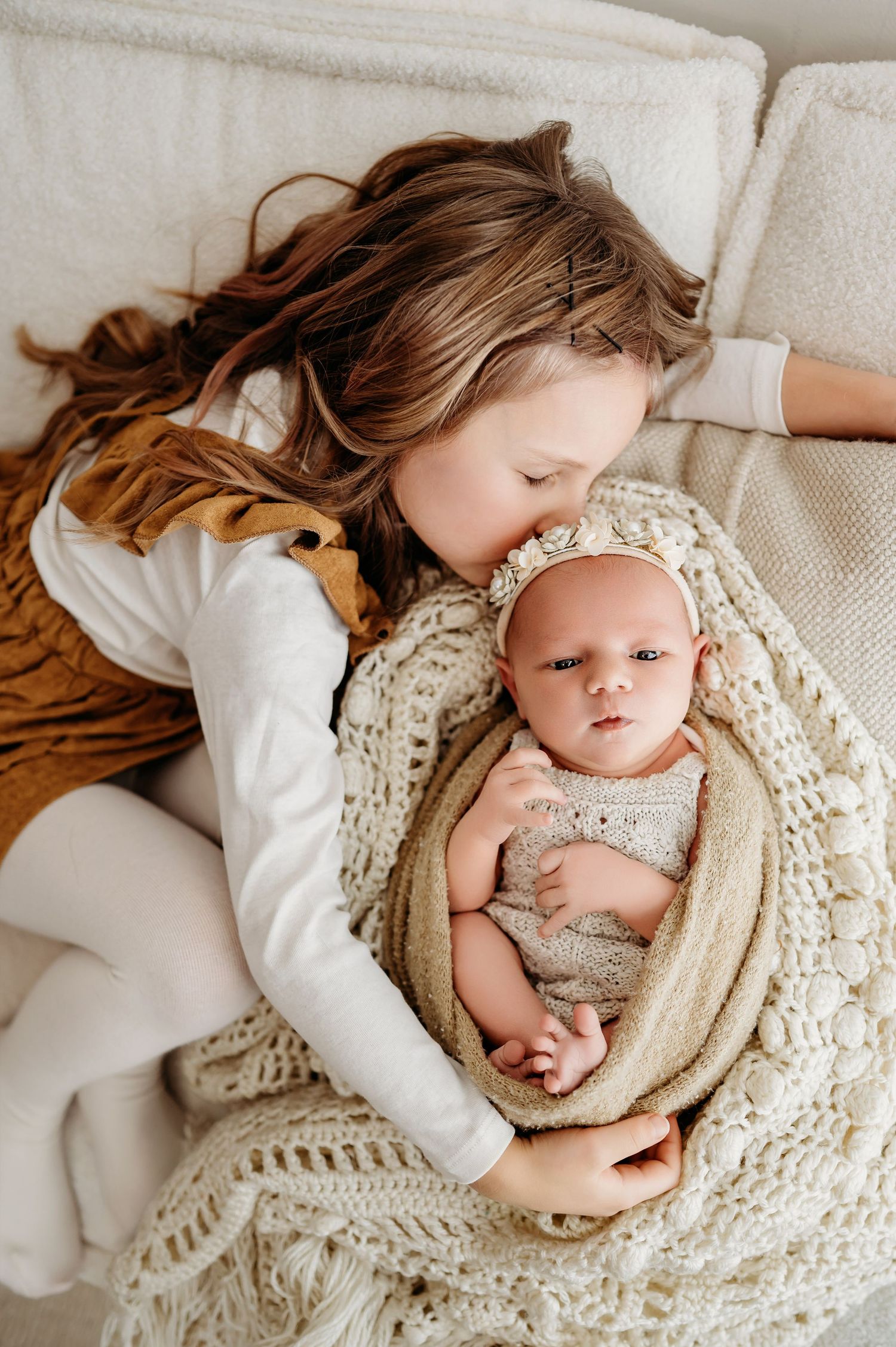 A tender moment as a young child in brown dress cuddles with a swaddled infant on a white couch with knit blanket.
