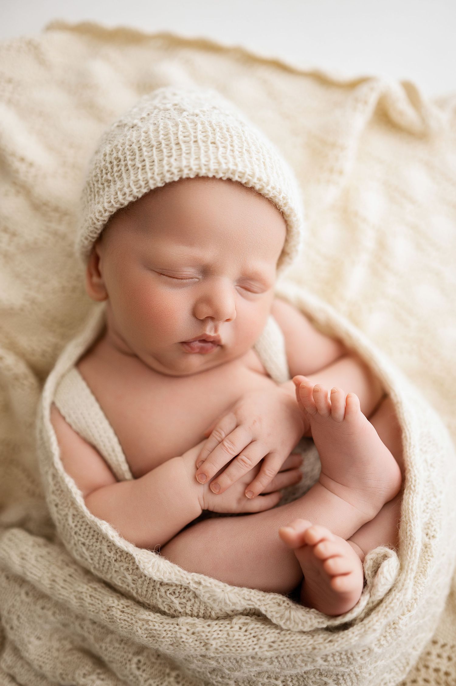 Sleeping newborn baby wrapped in soft cream knit blanket on neutral backdrop in peaceful curled pose.