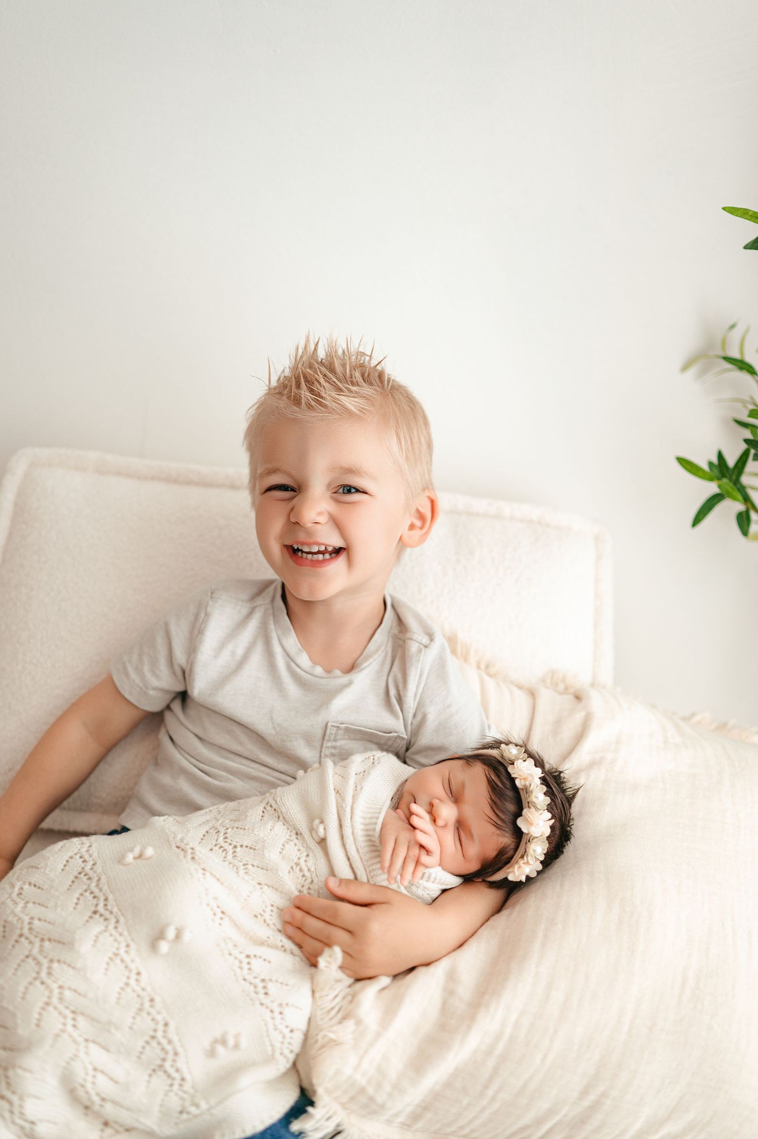 Two siblings share joyful moments together while laying on white bedding in a bright, airy room with plants.