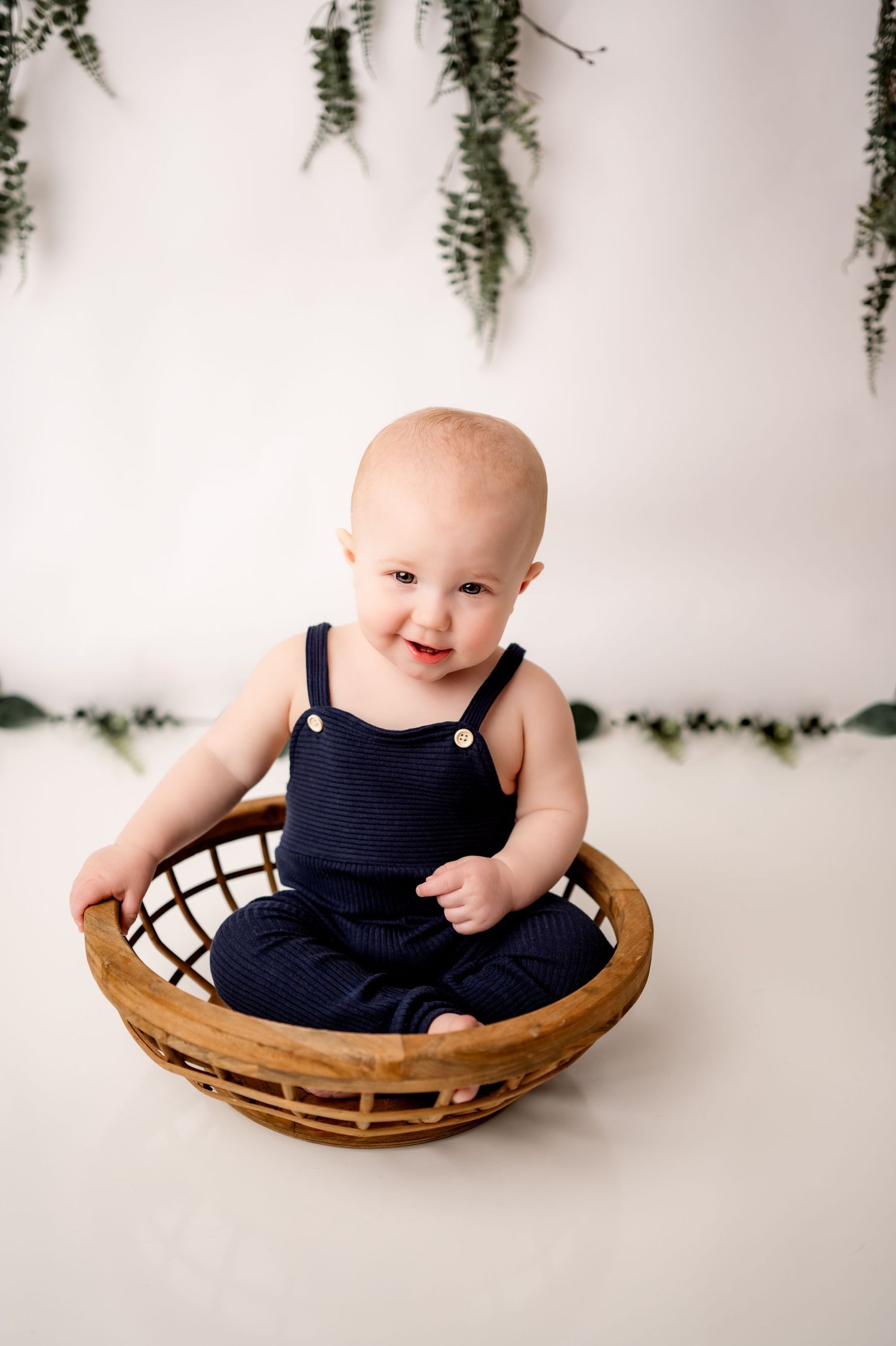 A baby wearing a navy dress sits in a curved wooden basket against a white backdrop with hanging greenery.