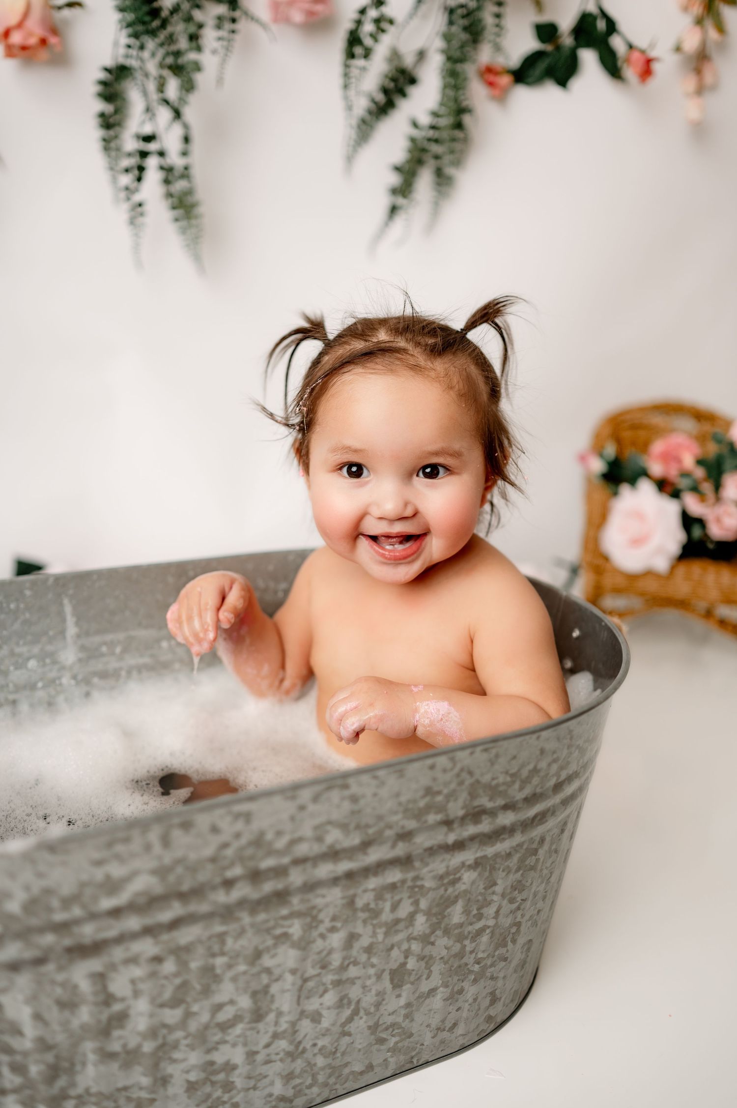 A happy baby takes a bubble bath in a vintage metal tub with floral decorations hanging on the wall behind.