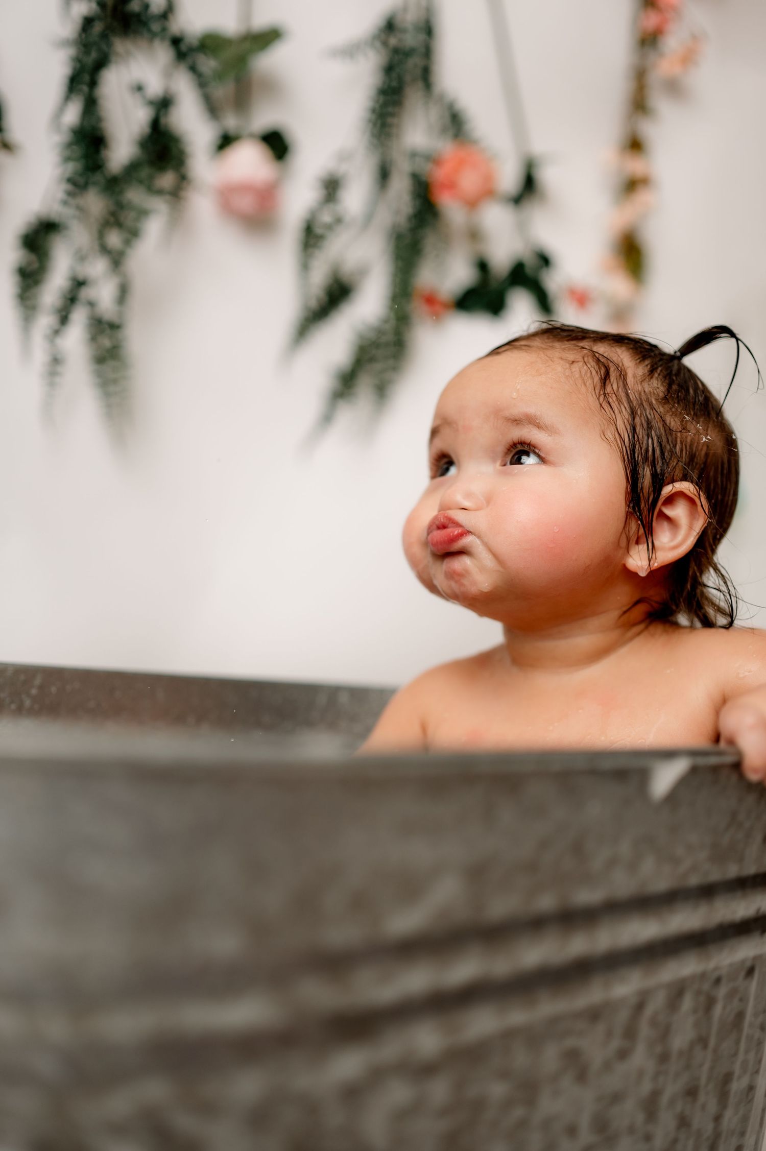 A small child peeks over the edge of a gray bathtub with floral decorations hanging on the wall in the background.