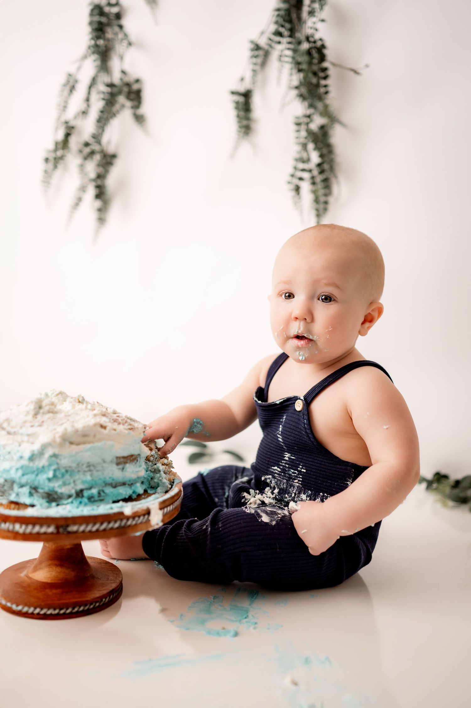 Baby in navy outfit explores a blue and white smash cake during a studio photography session with hanging ferns.