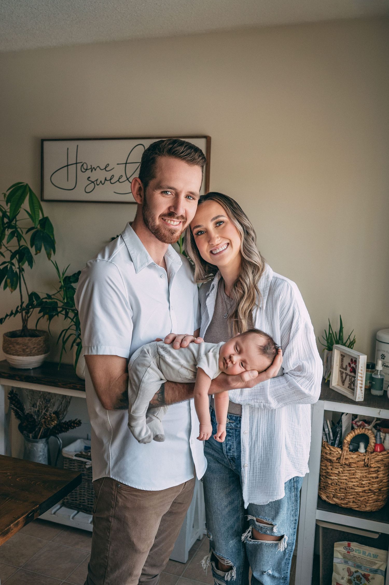 Young couple standing in home interior cradling their sleeping newborn baby against a wall with decor.