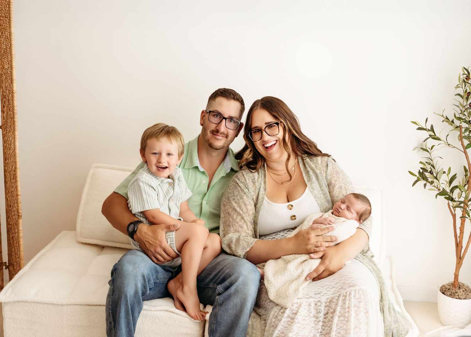 A bright and airy studio photography session shows a family lounging together on a white sofa against neutral walls.