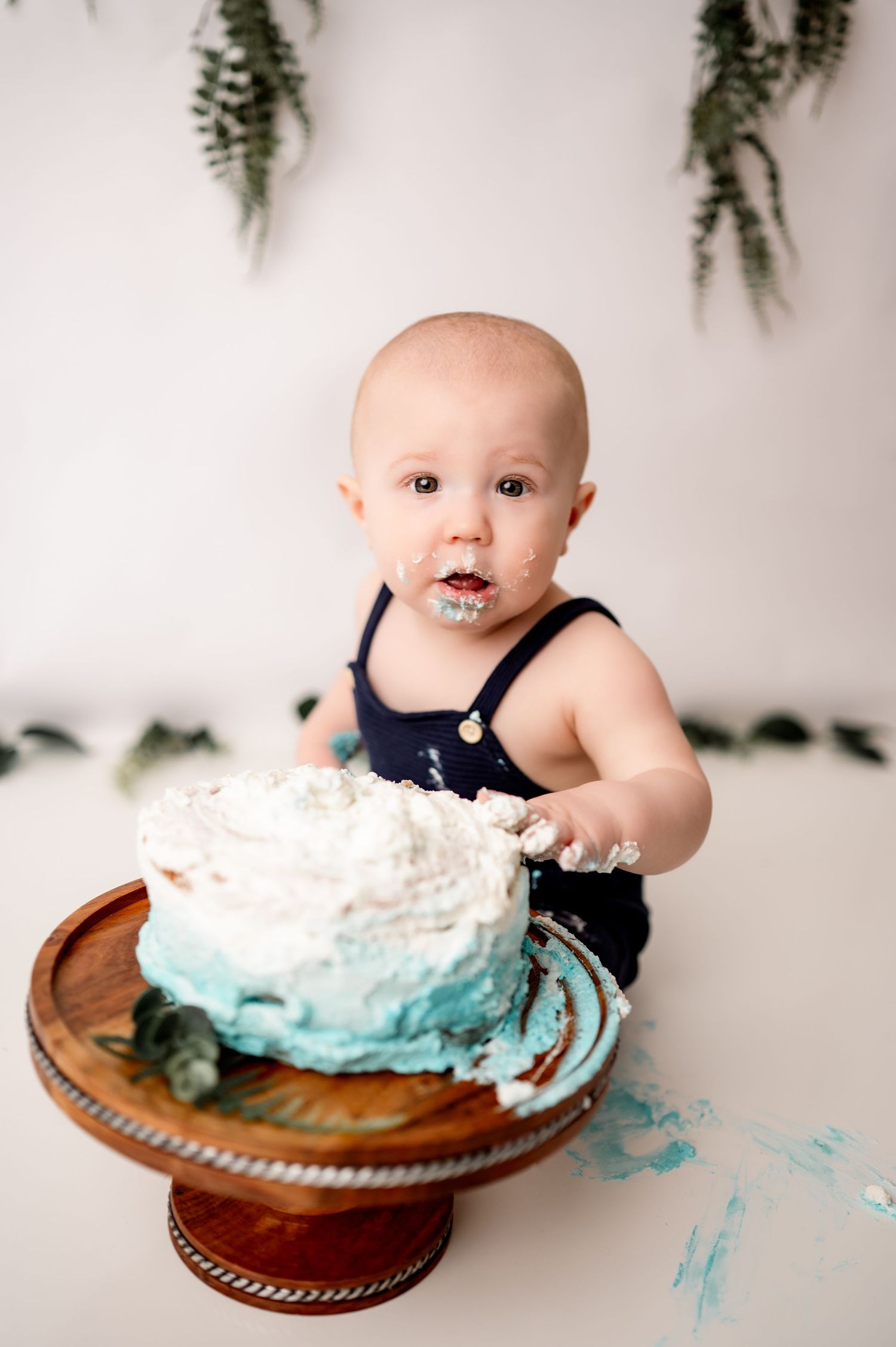 Baby in navy blue outfit dives face-first into smash cake with turquoise and white frosting on wooden plate.