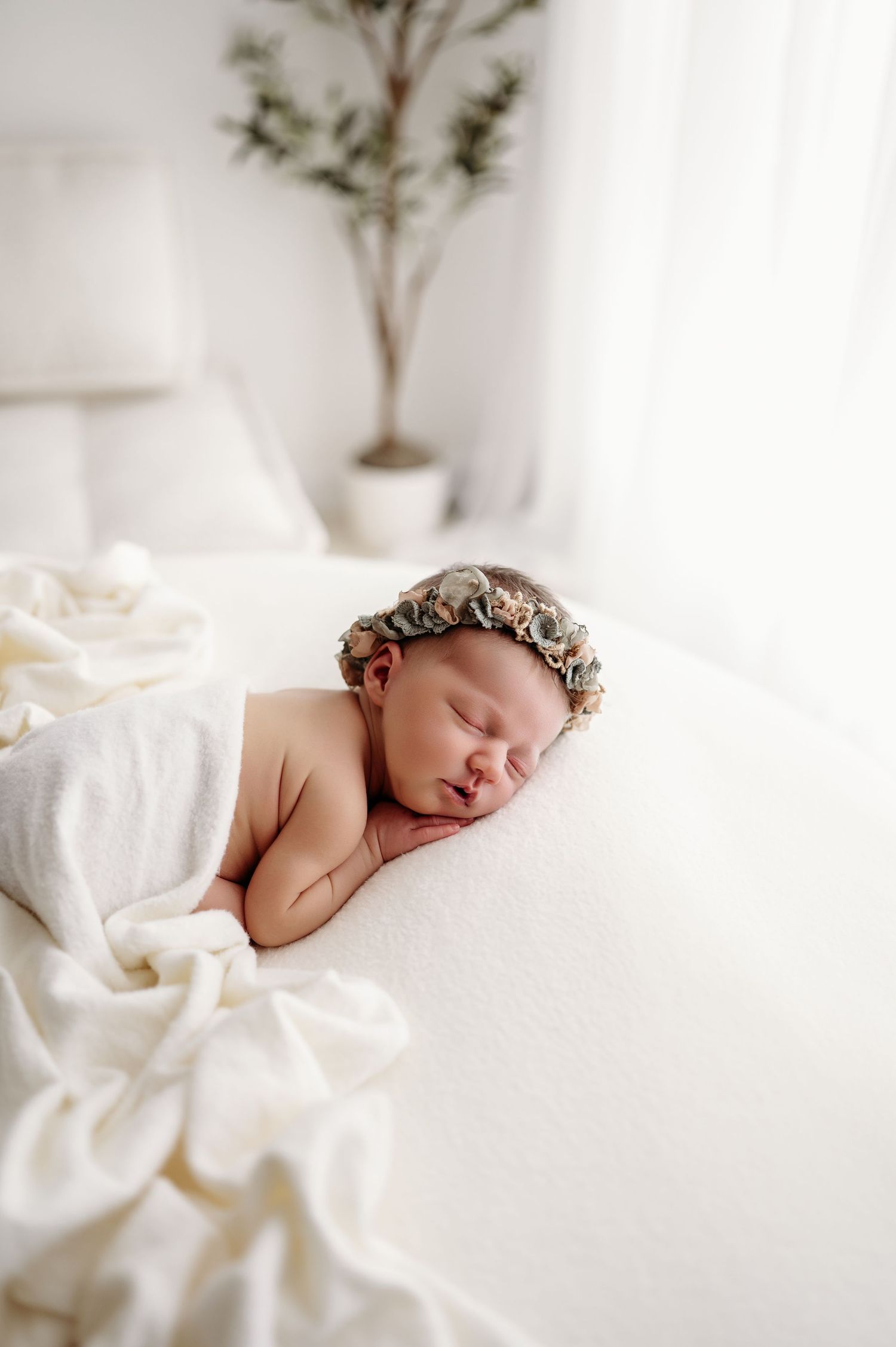 Sleeping newborn wearing floral crown lies peacefully on soft white bedding with minimal plant decor in background during a studio session w