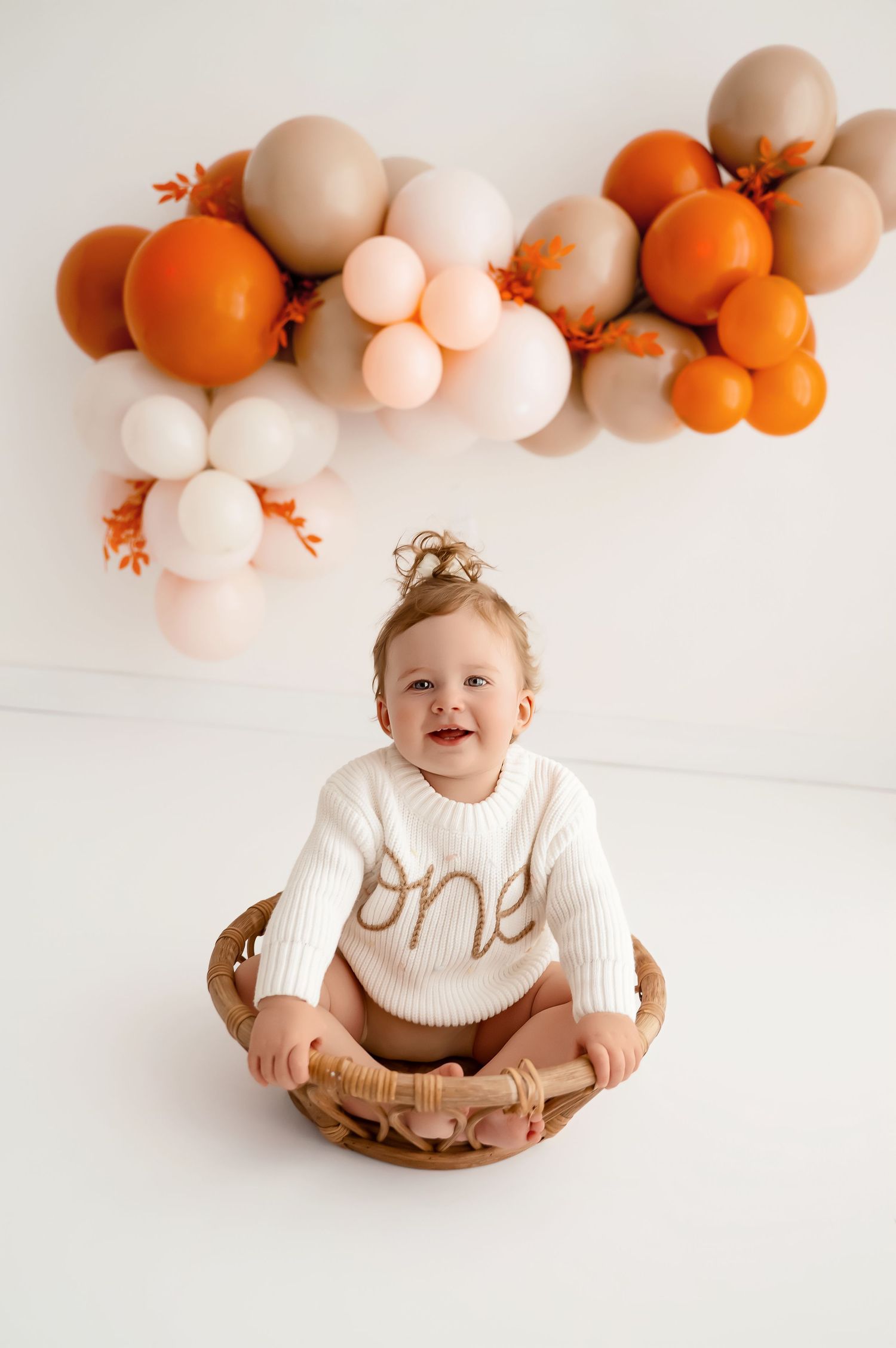 First birthday photo session featuring white and orange balloon decor in a clean, modern setting.