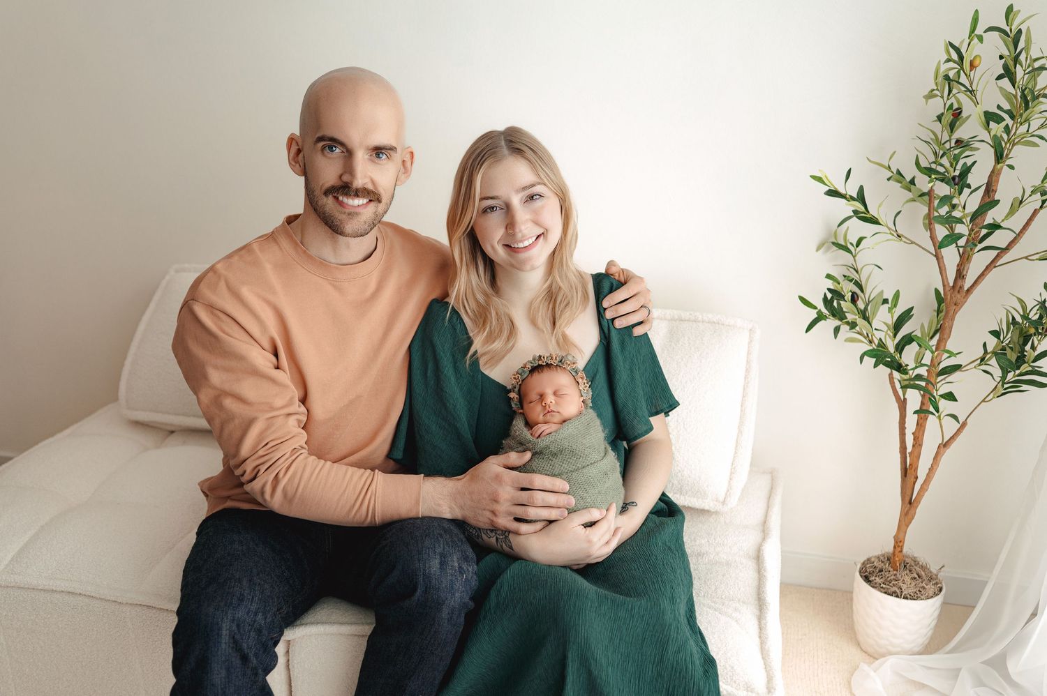 A couple sits on a beige couch in a bright minimalist room with a small potted tree in the corner.