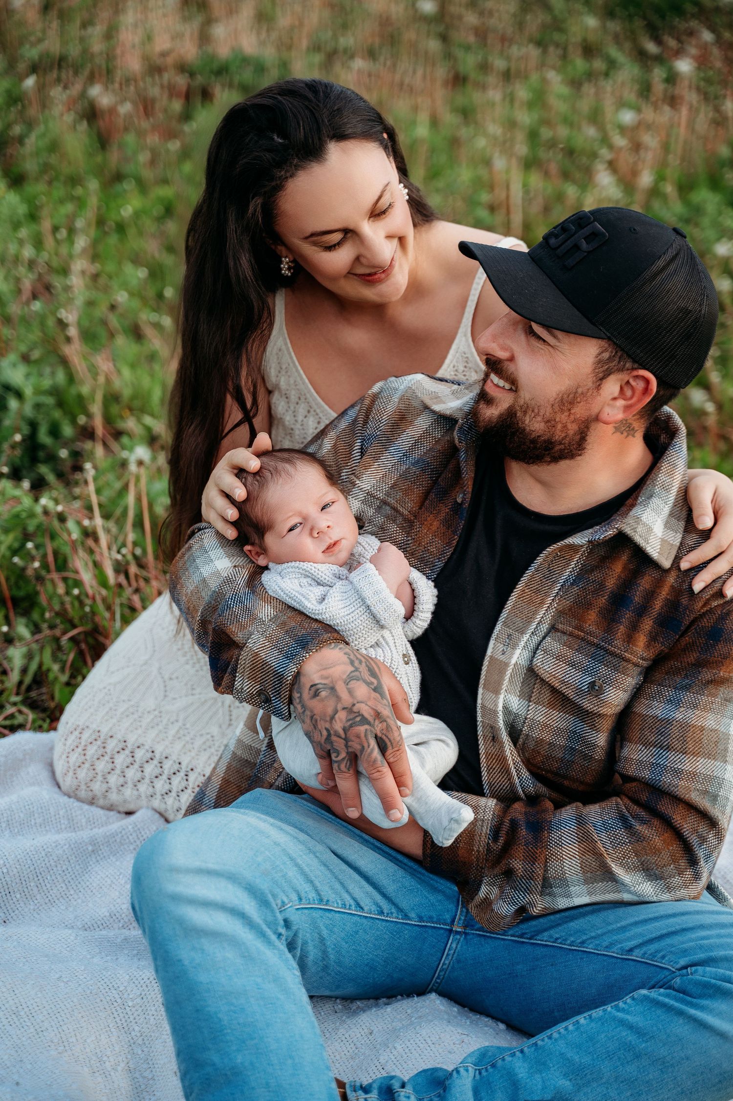 Family of three sitting together on a blanket outdoors, sharing tender moments in matching plaid shirts.