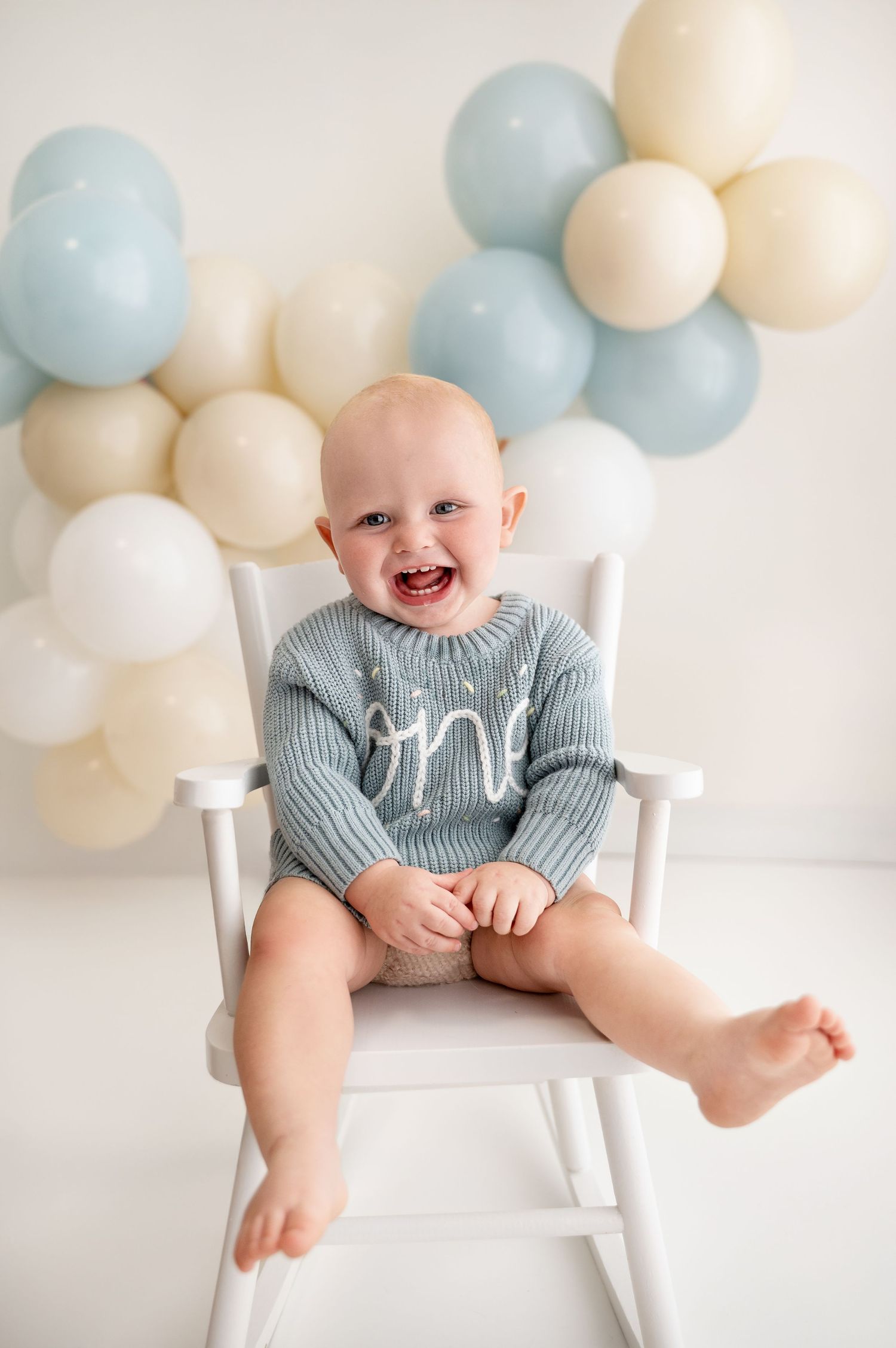 Baby wearing gray sweater sits in white chair against pastel balloon backdrop.