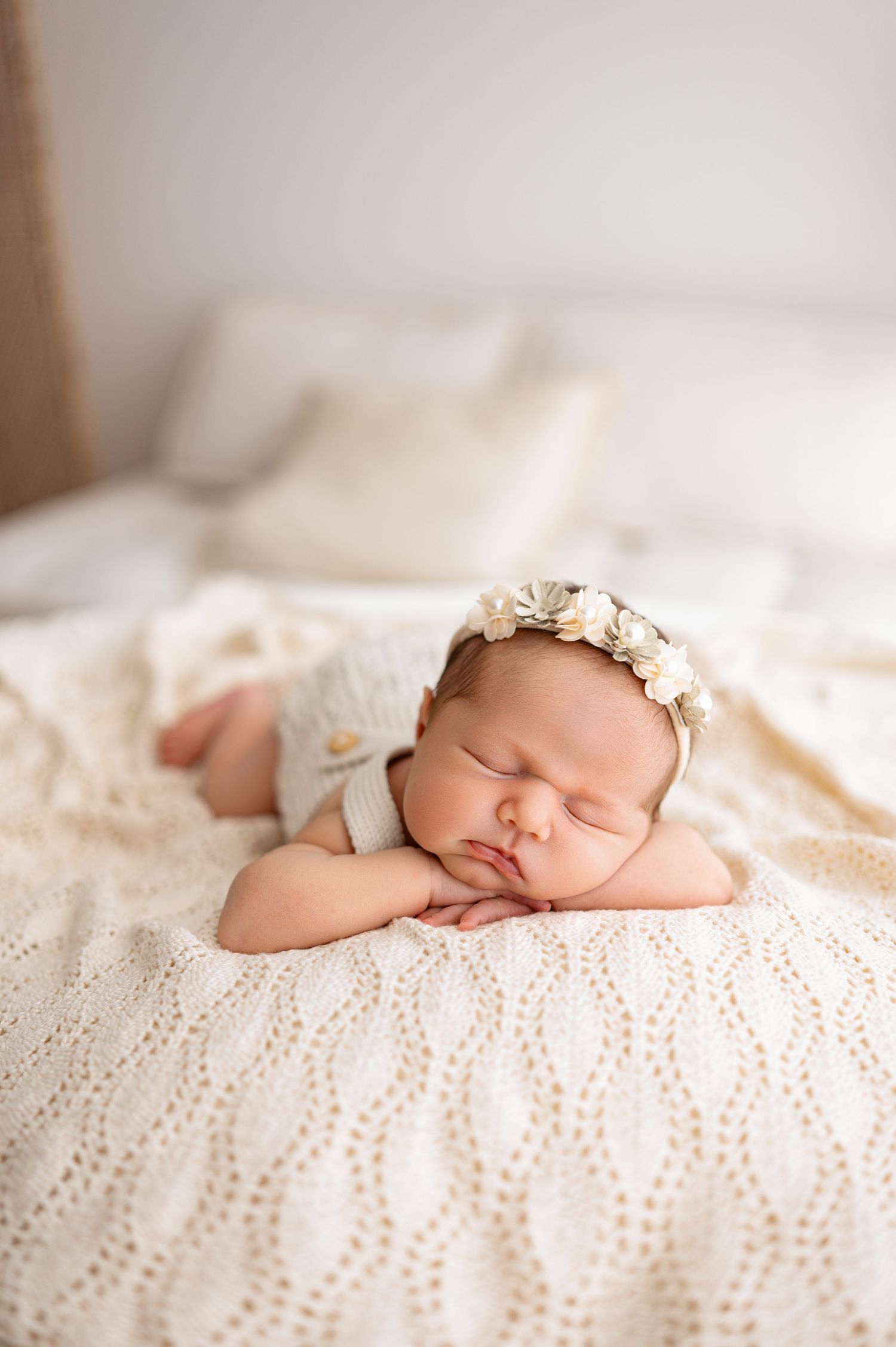 A sleeping newborn rests peacefully on white lace blankets wearing a delicate floral headband.