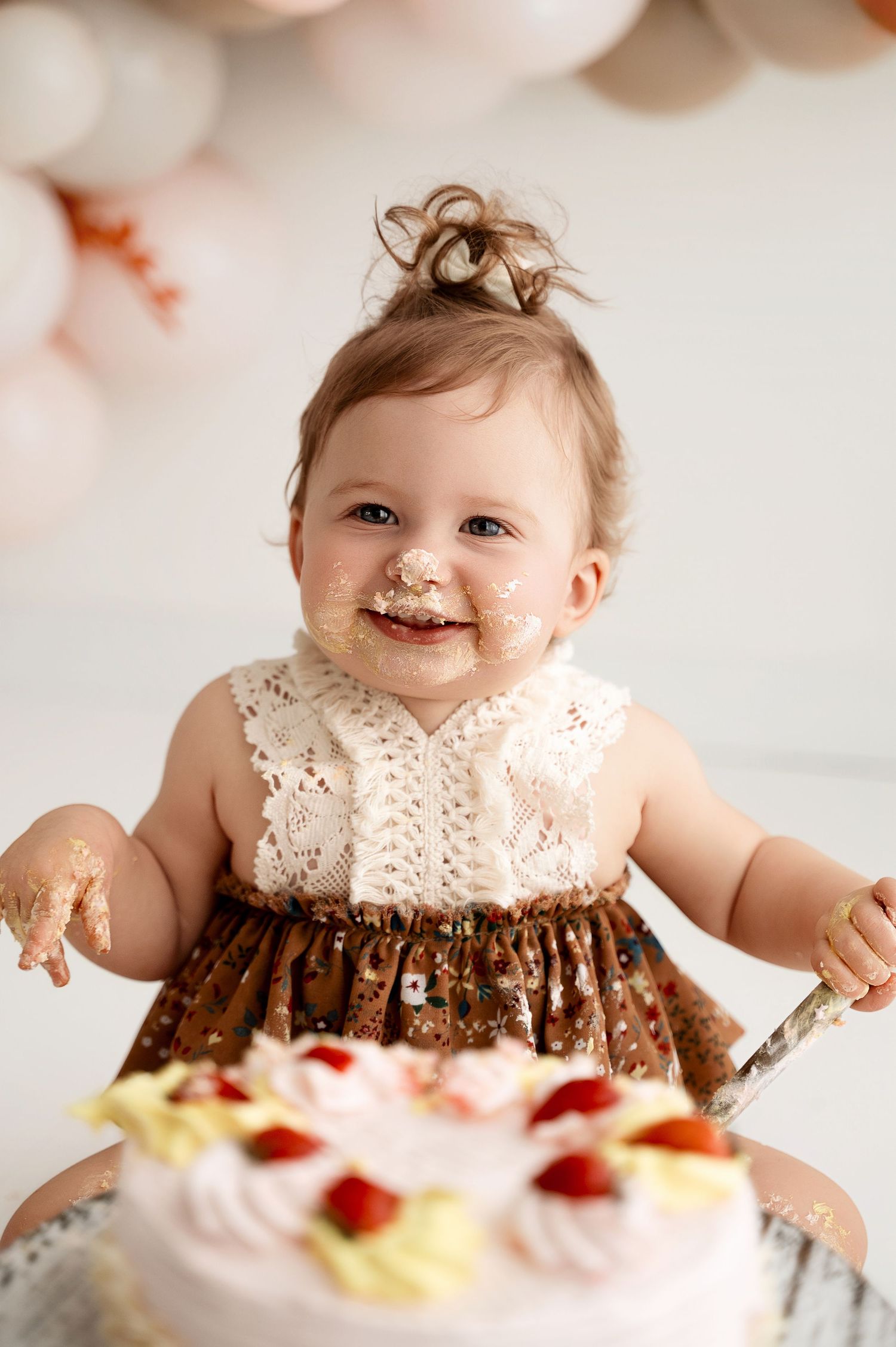 Baby wearing lace dress sits in front of white and peach balloon garland during cake smash photo session.