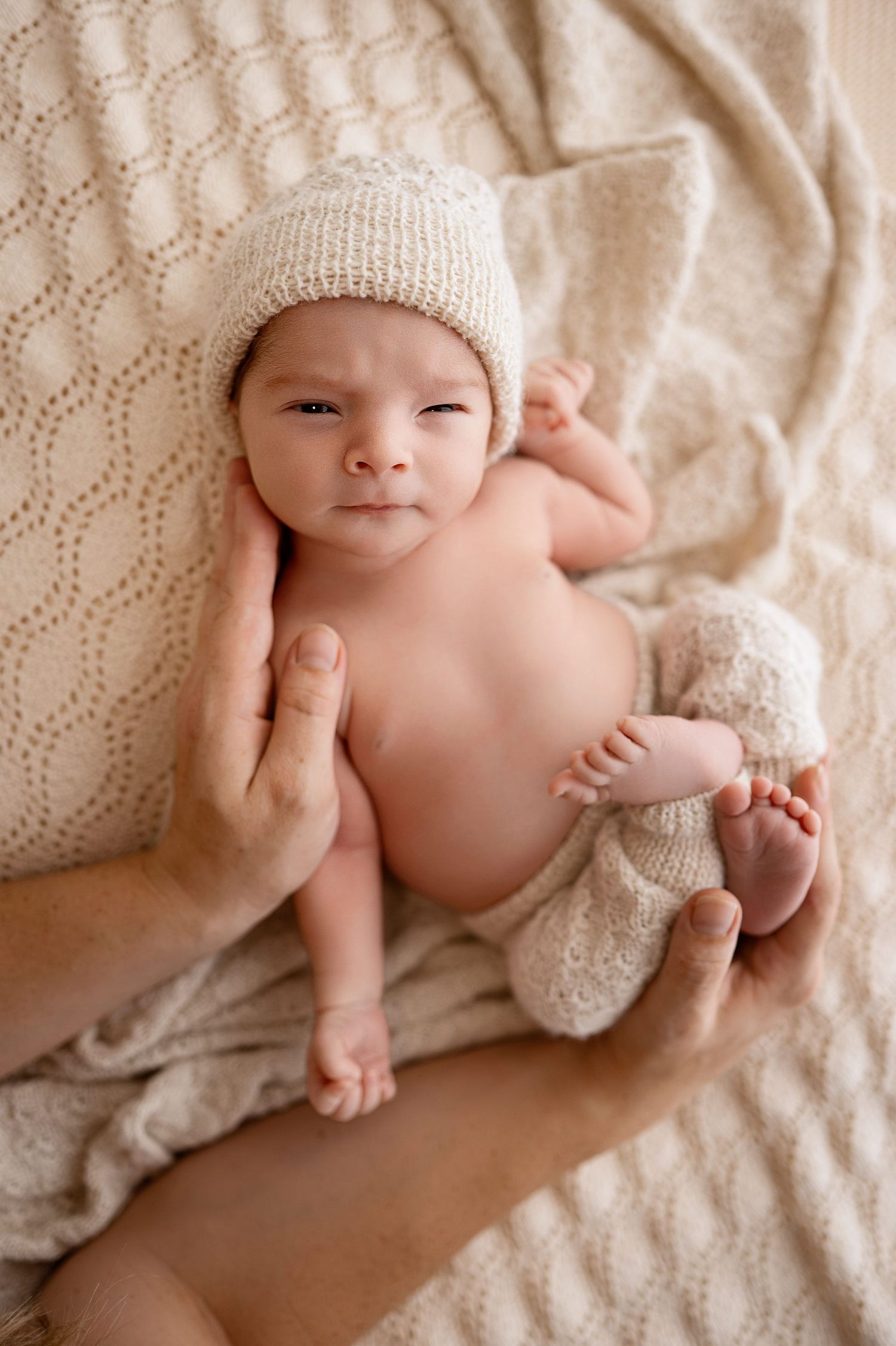 Newborn baby in white knit hat lying on textured cream blanket during professional photo session.