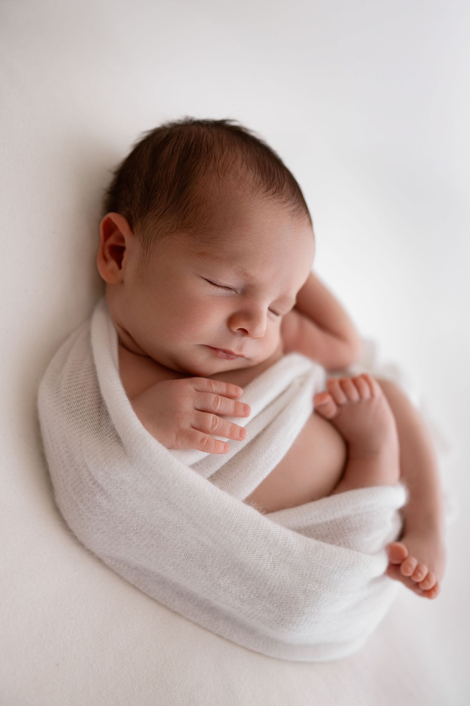 Peaceful sleeping newborn baby wrapped in soft white blanket against light background.