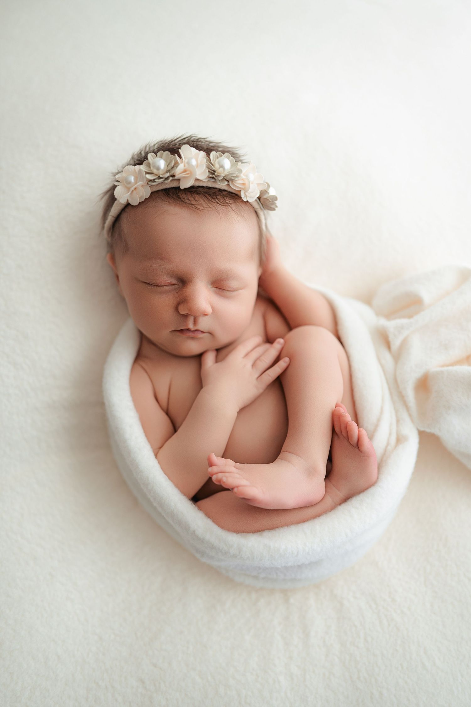Sleeping newborn wrapped in white blankets with floral headpiece curled up in curved moon-shaped prop for professional photo.