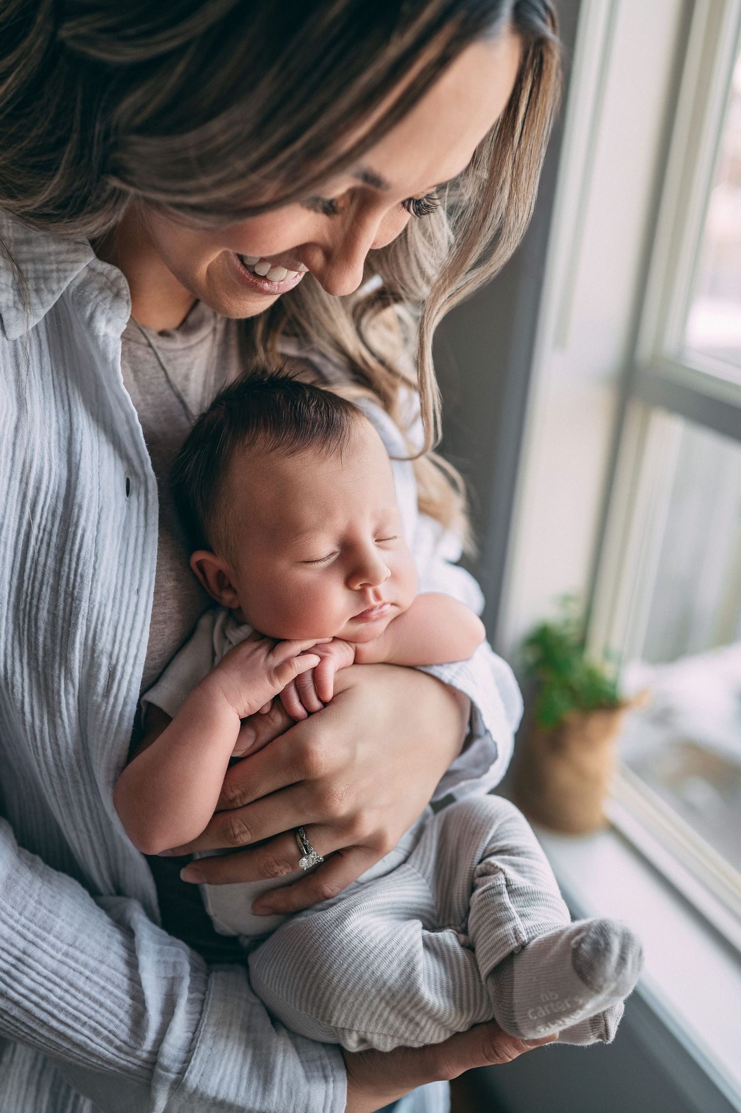 A peaceful moment by the window as a sleeping newborn baby rests in loving arms wearing soft gray clothing.