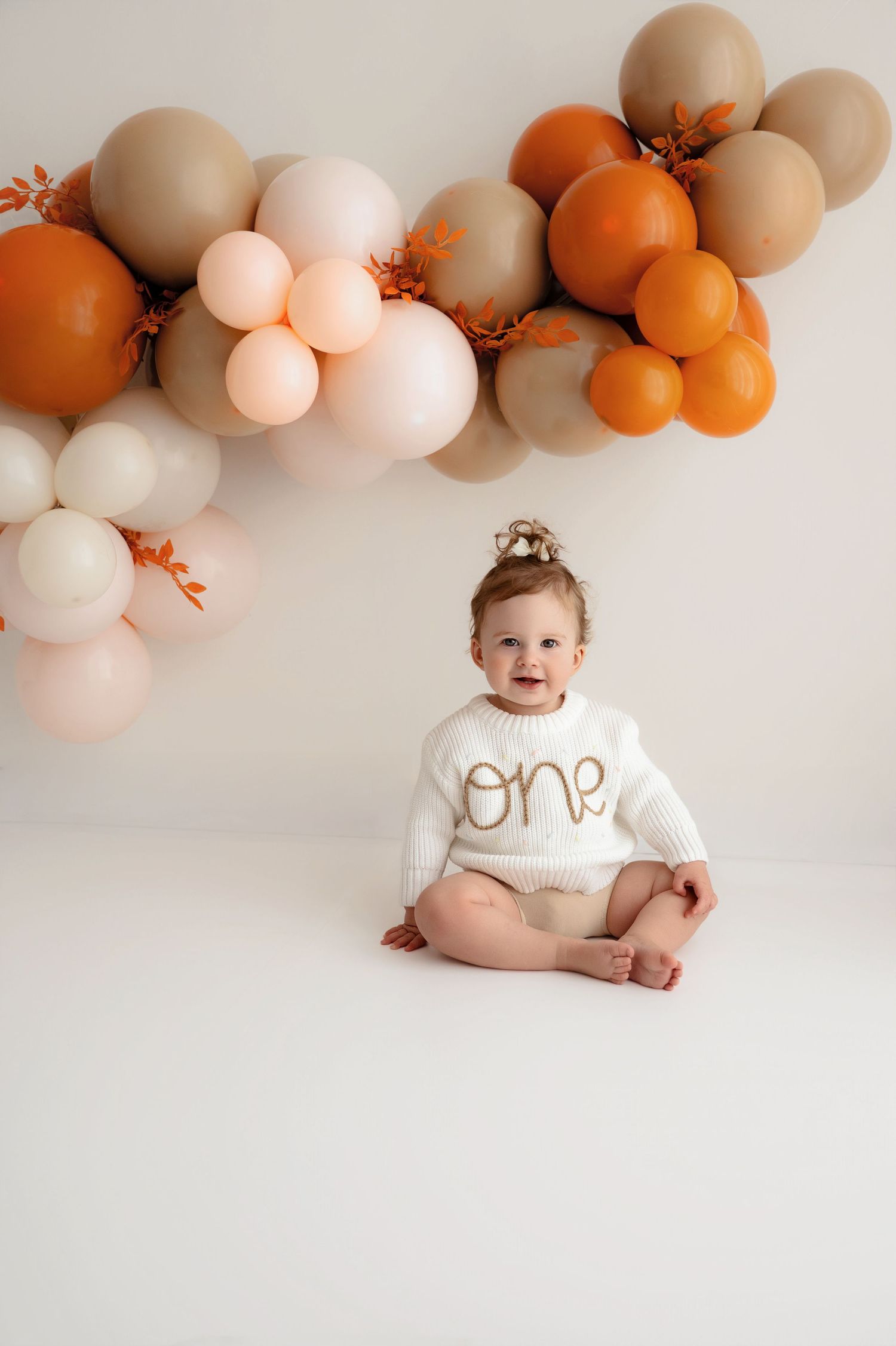 Baby milestone photo session with peach and orange balloon arch against white backdrop featuring one year old in white outfit.