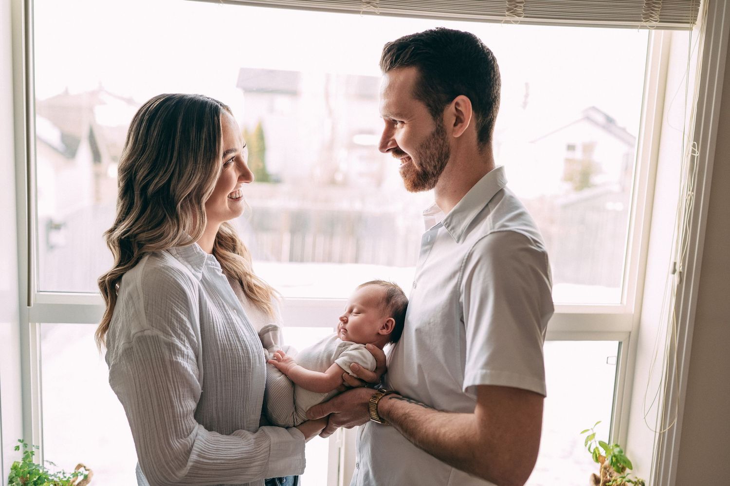 A couple in white shirts tenderly holds their newborn baby near a bright window at home.