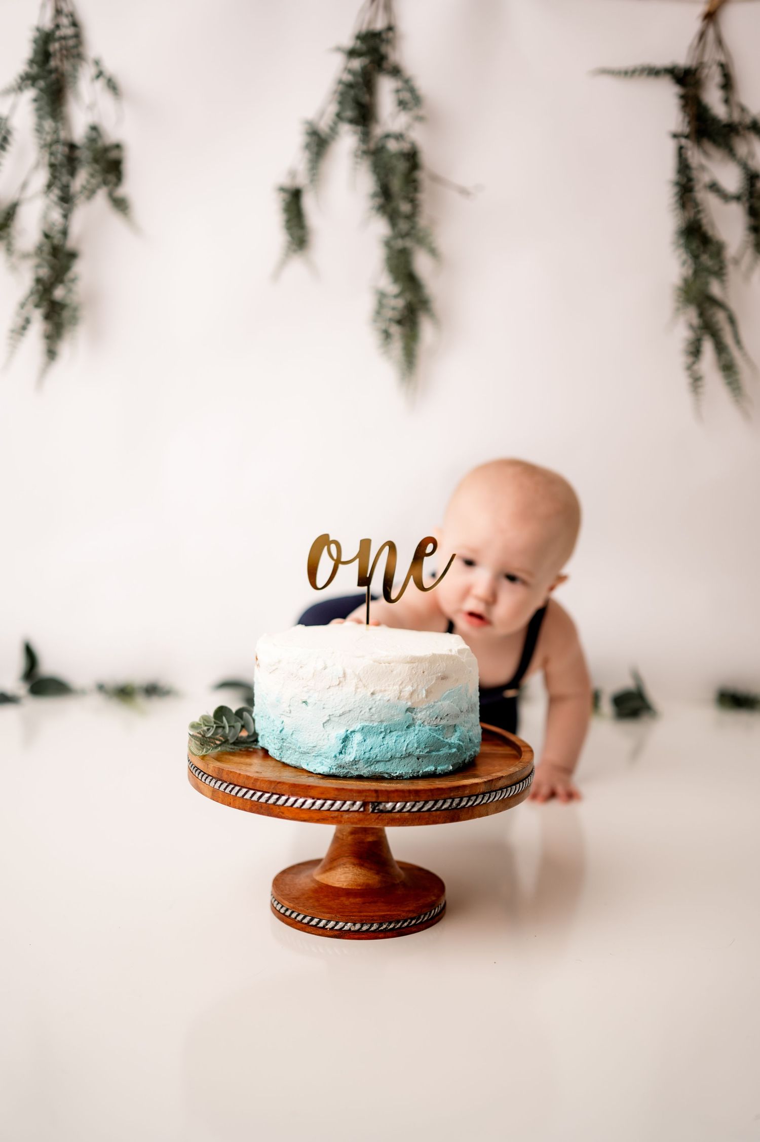 A cake smash photo setup with turquoise cake on wooden stand and greenery decorations in the background.