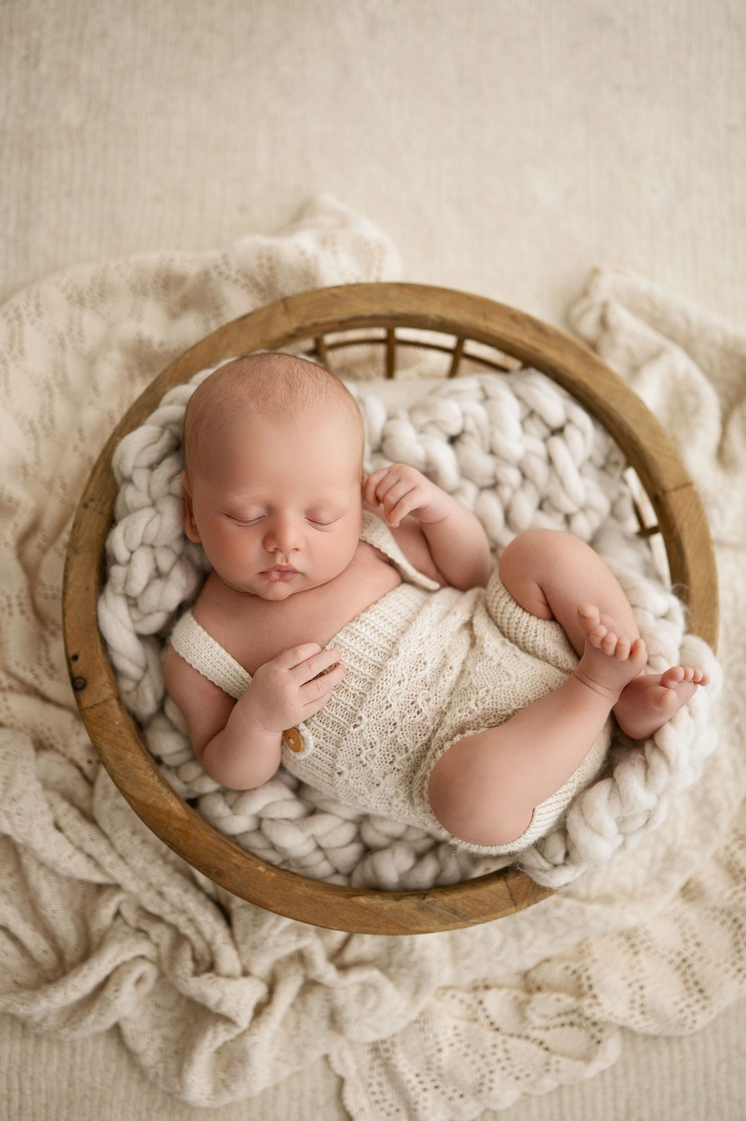 A sleeping newborn nestled in a wooden bowl with soft white textiles and delicate lace details.