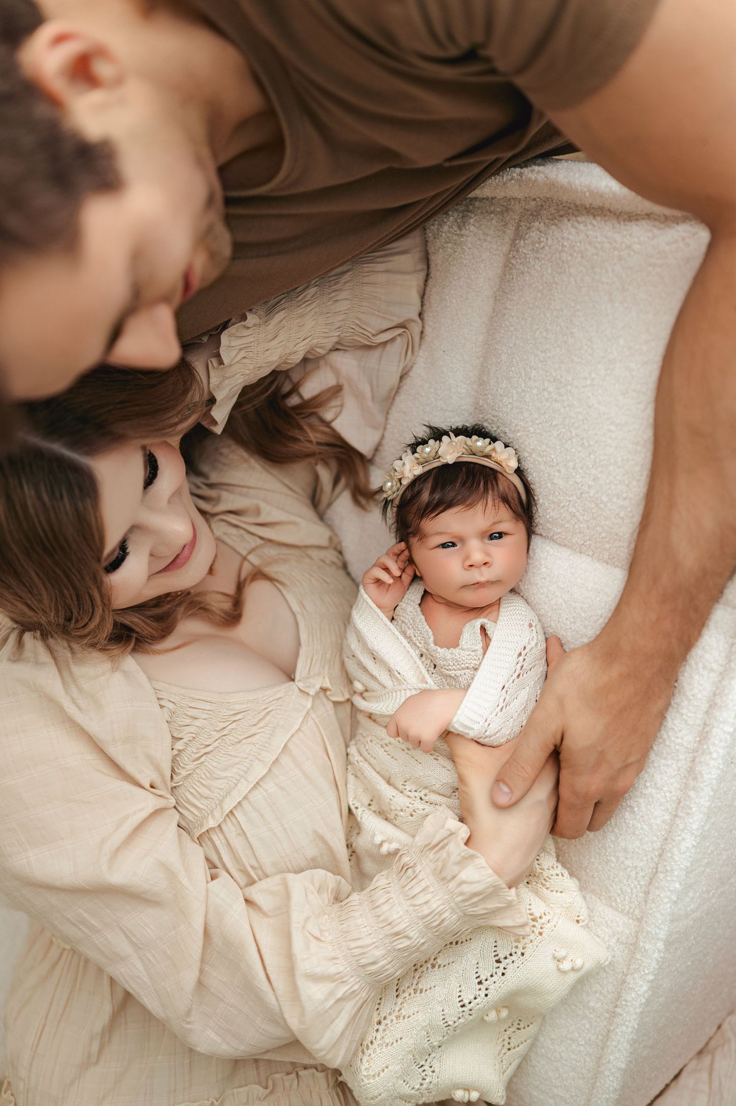 A family lies together on white bedding in soft natural light wearing cream colored clothing during an intimate portrait session.