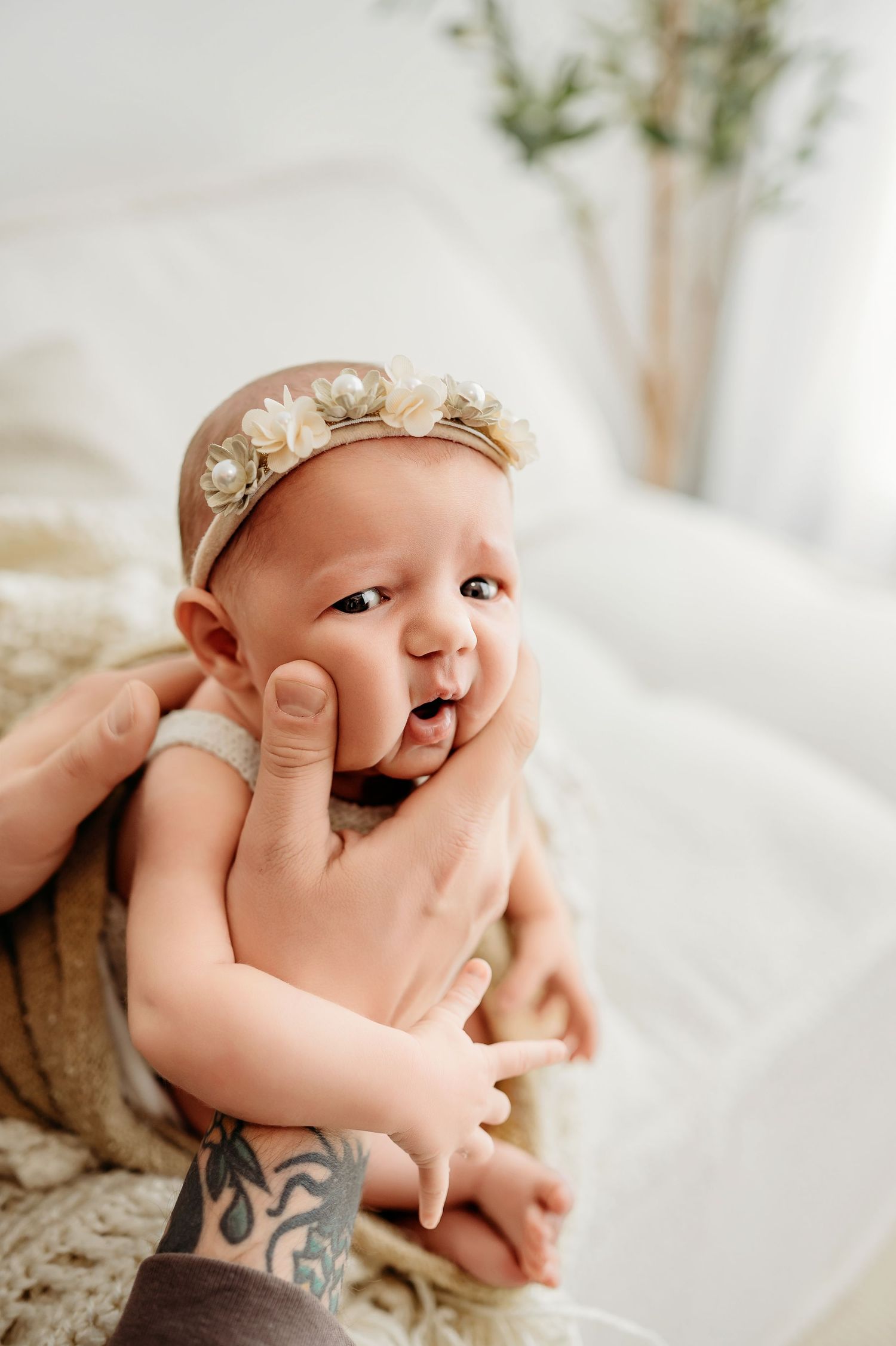 A baby wearing a delicate floral headband smiles while being gently held against light-colored bedding.