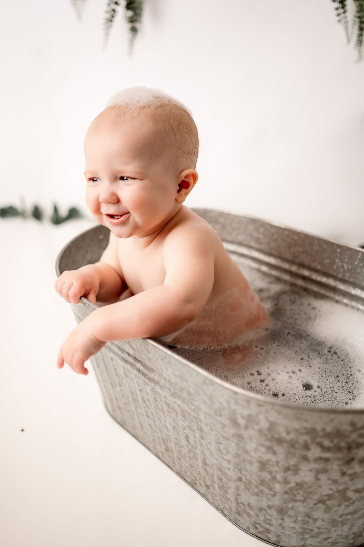 A happy baby enjoys bathtime in a gray tub filled with soapy bubbles and water.