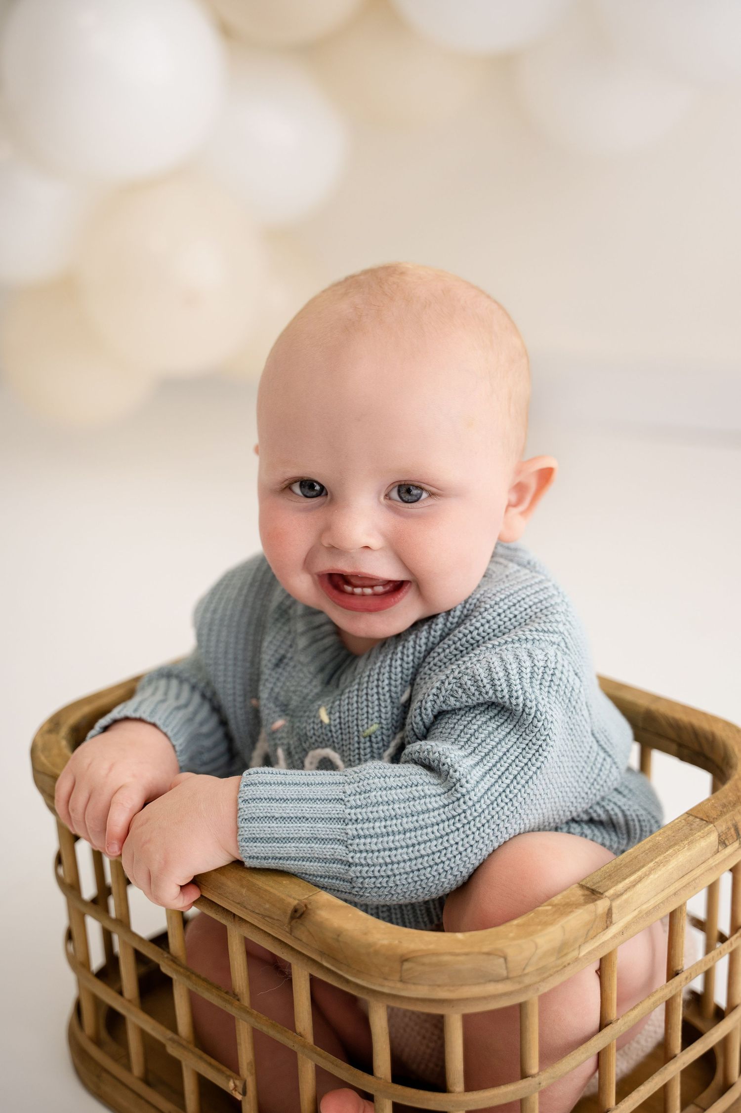 Baby in a gray knit sweater smiling while laying in a wooden basket against a light background.