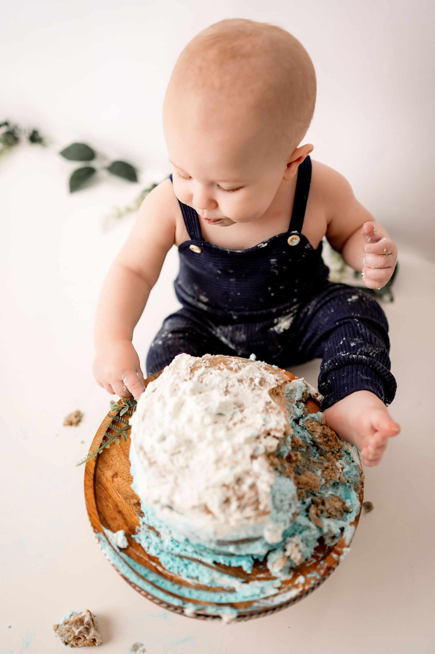Baby in navy outfit explores and plays with a smashed blue and white cake during a cake smash photo session.