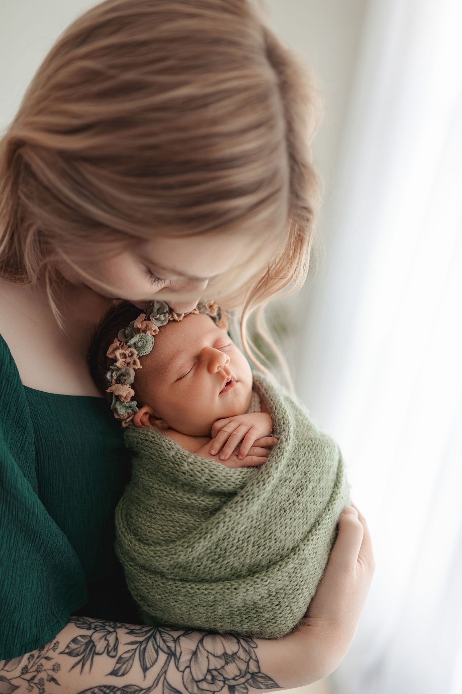 Newborn baby wrapped in green knit blanket with flower crown laying peacefully during professional photo shoot.