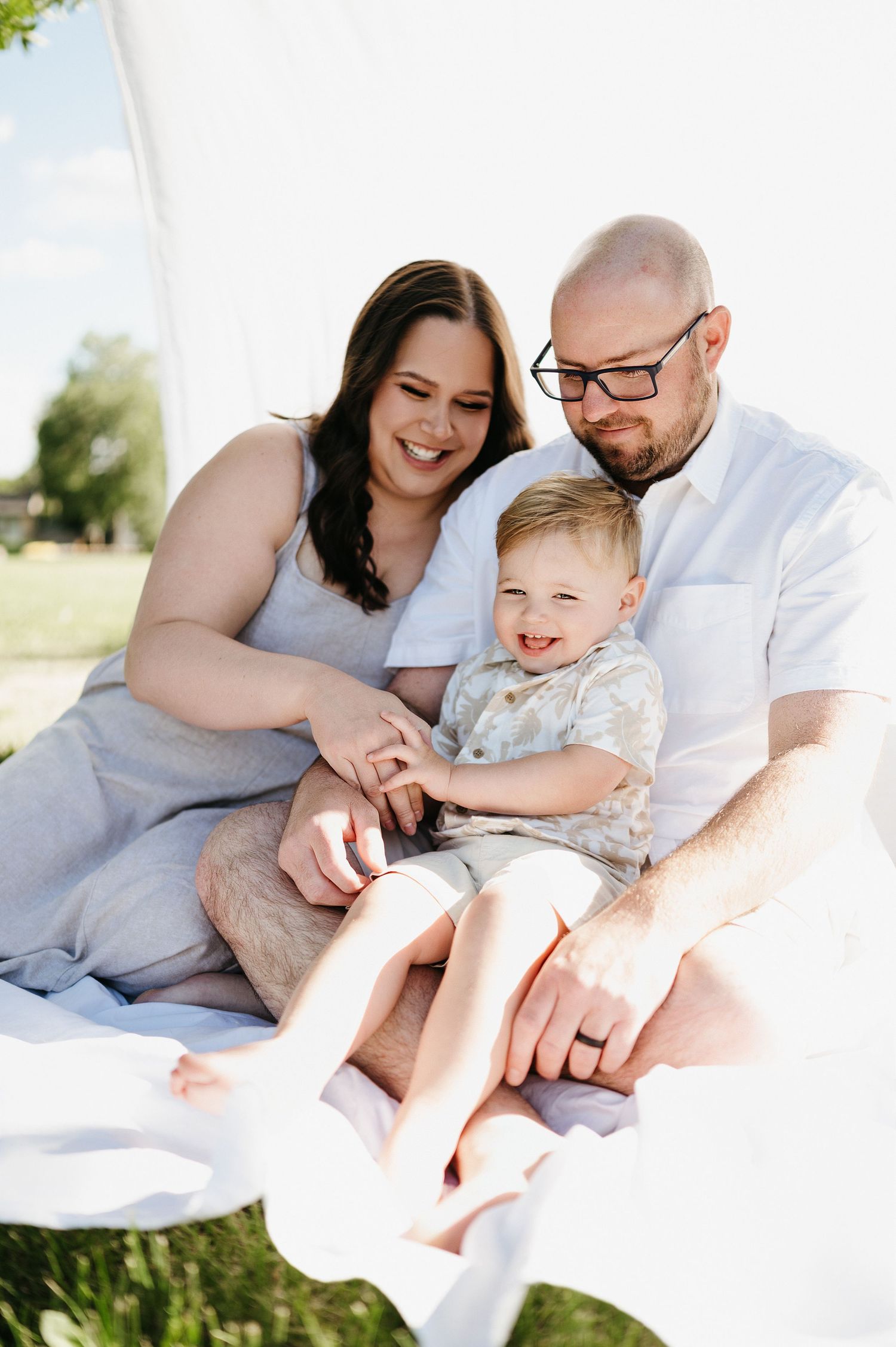 Family cuddles together on white blanket outdoors in sunlit field wearing casual summer clothing.