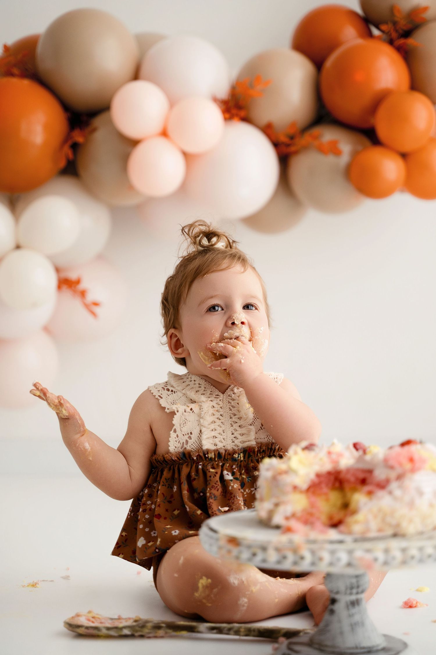 Baby in brown dress enjoying first birthday cake smash session with orange and white balloon backdrop.