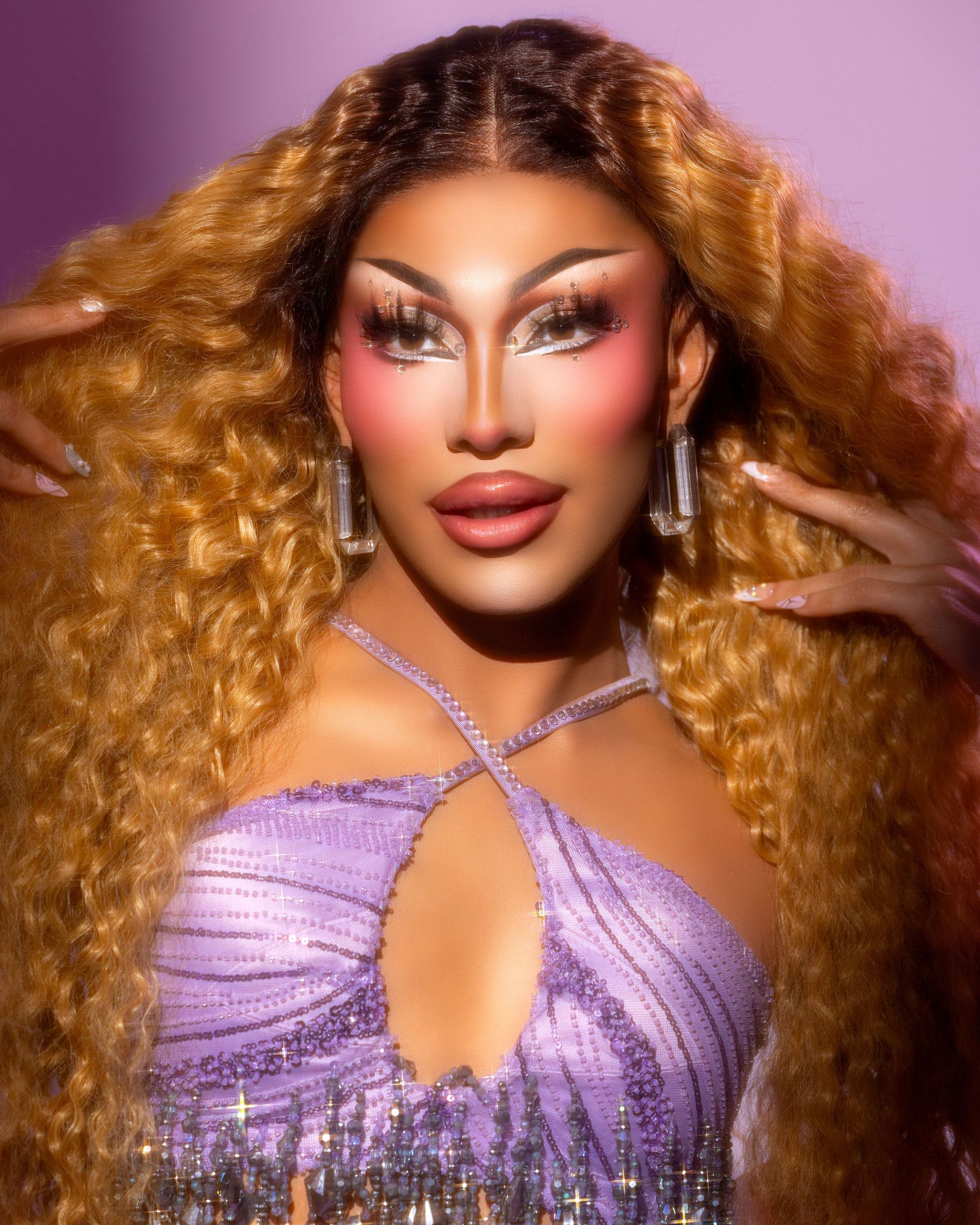 Drag performer wearing a sparkly purple dress with long honey-blonde curly hair and dramatic makeup.