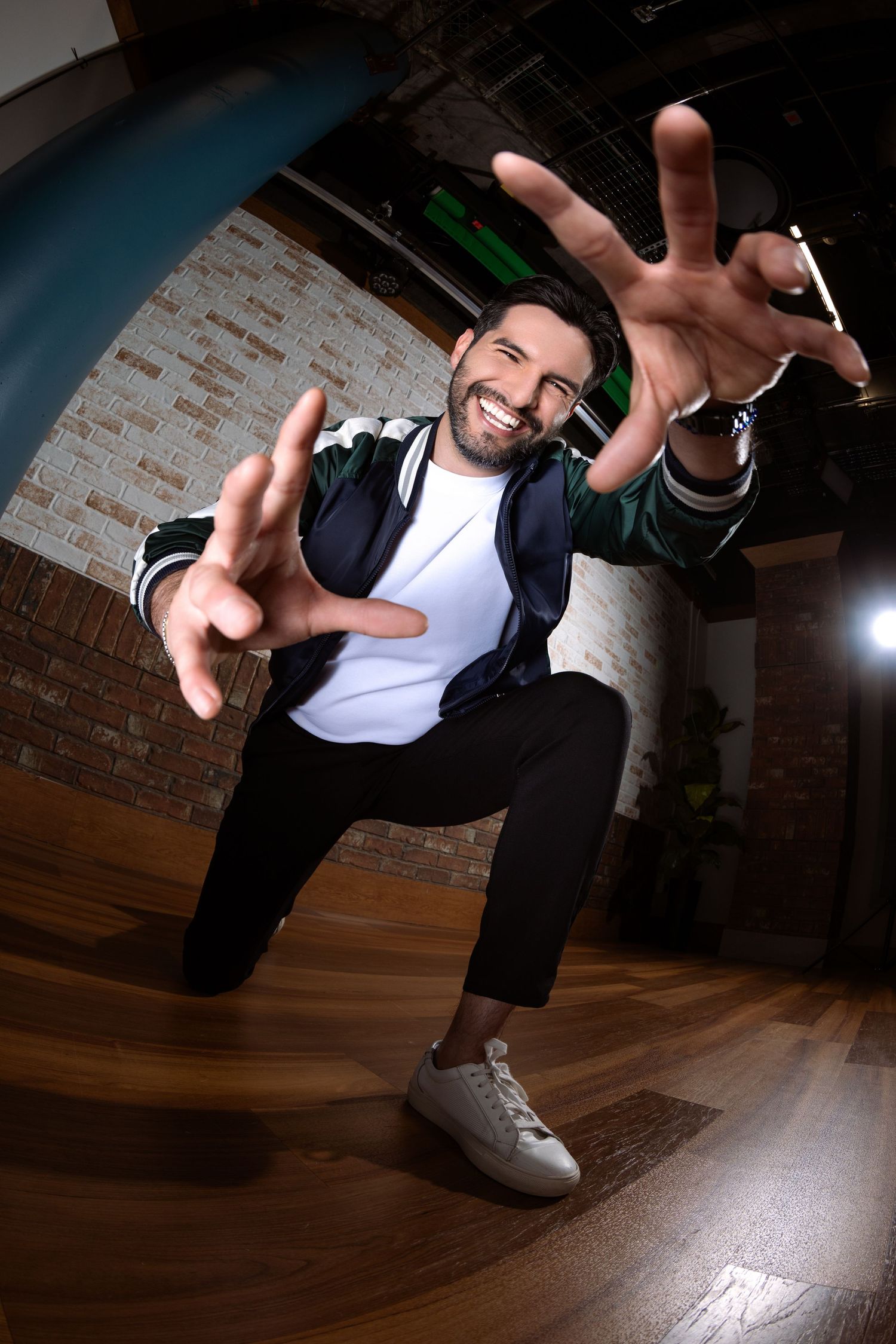 Smiling man in jacket reaching toward camera with both hands in a dynamic fisheye lens pose on wooden floor.