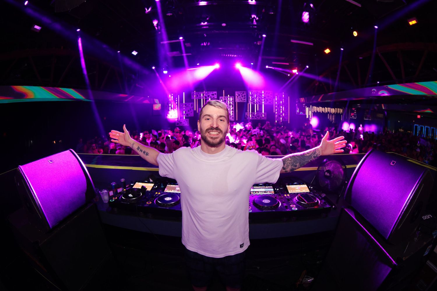 DJ performs behind booth at nightclub with purple lighting and raised arms against vibrant dance floor atmosphere.