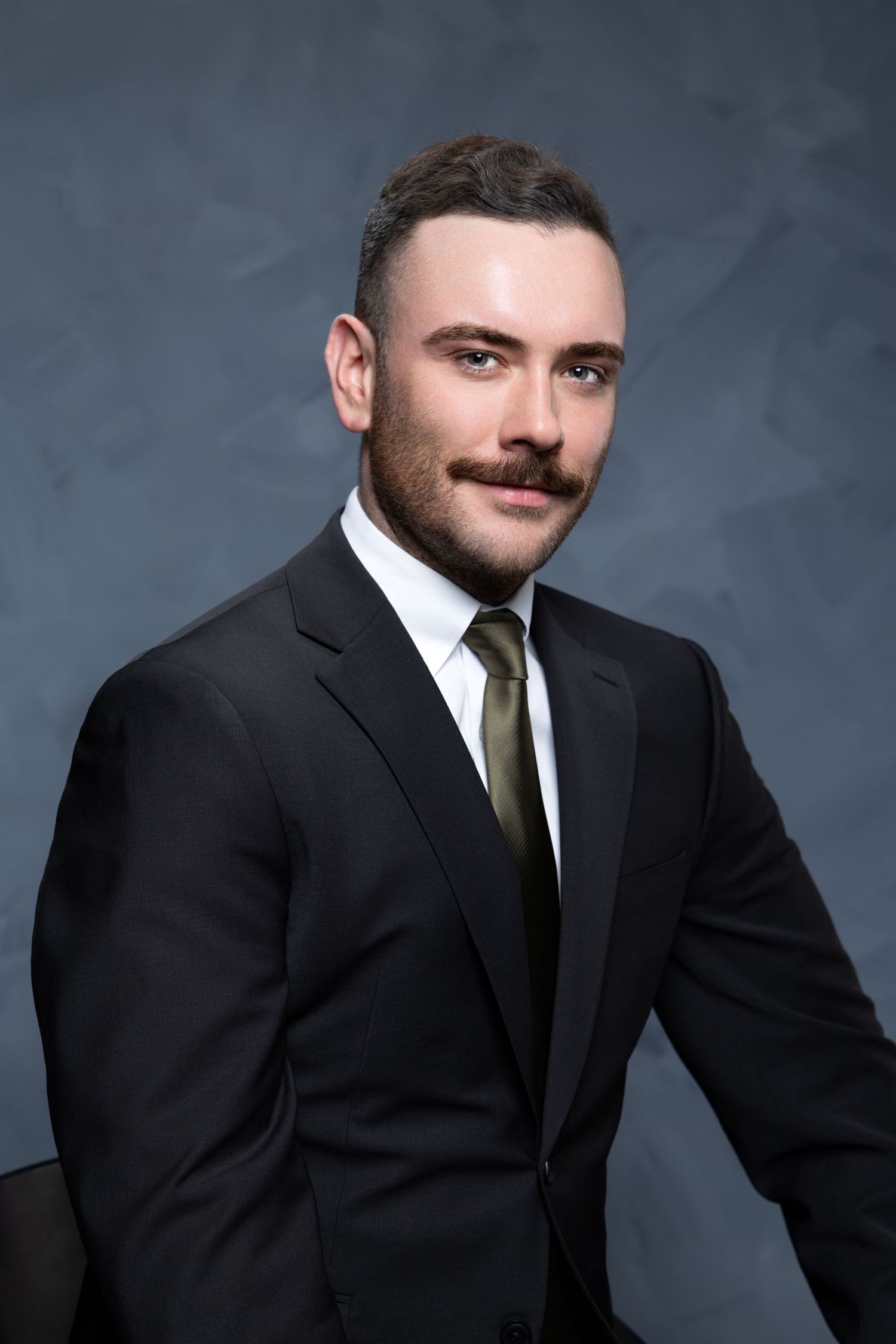Professional headshot series of a person in a black suit and tie against a dark gray studio backdrop.