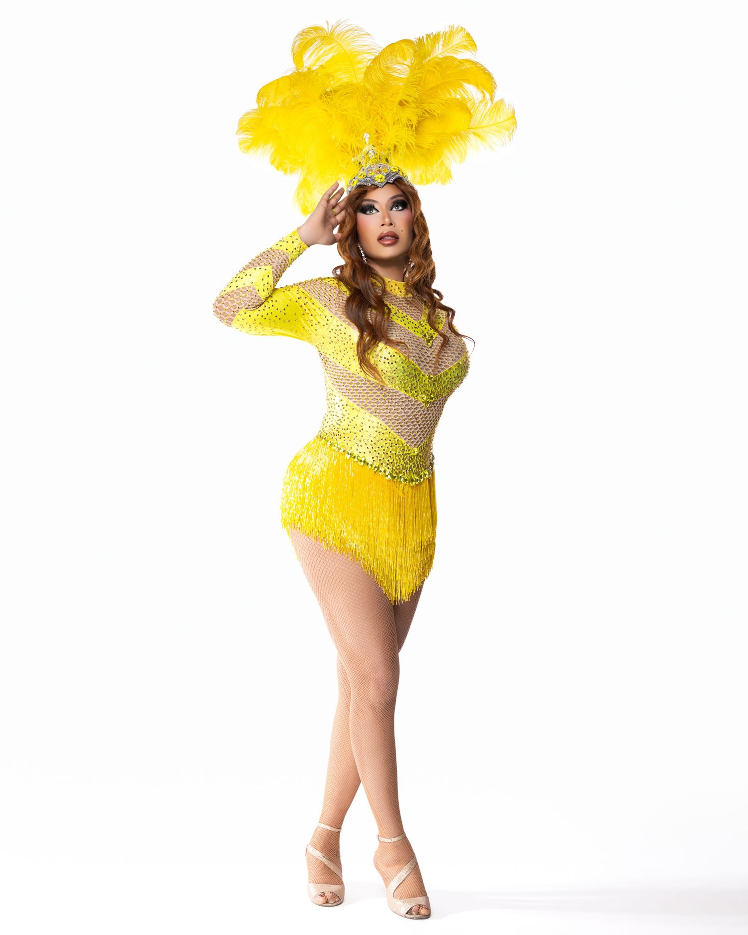 Performer in yellow sequined fringe costume with feathered headdress and crown poses on white background.