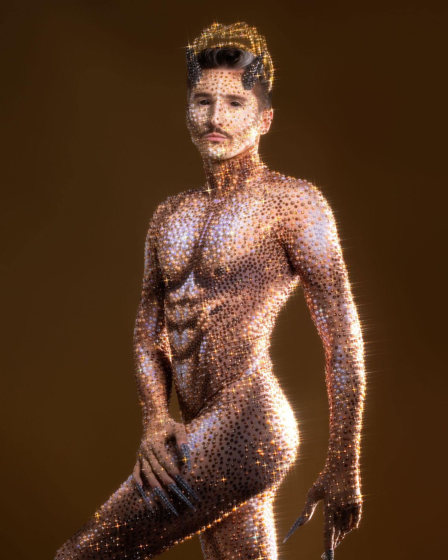 A muscular man covered in glittering gold body paint poses dramatically against a warm brown background.
