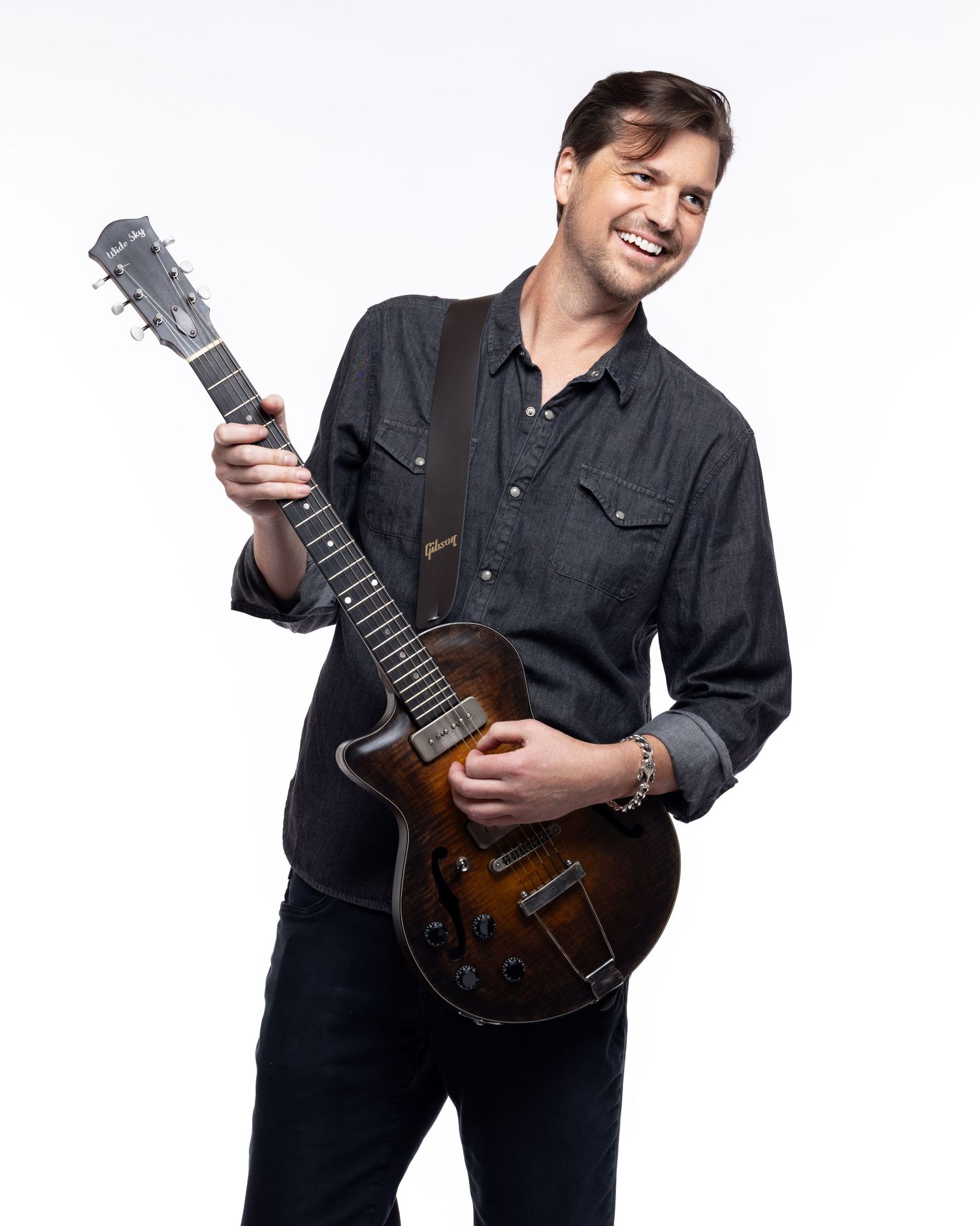 Musician in black button-down shirt holds and poses with a brown electric guitar against white background.