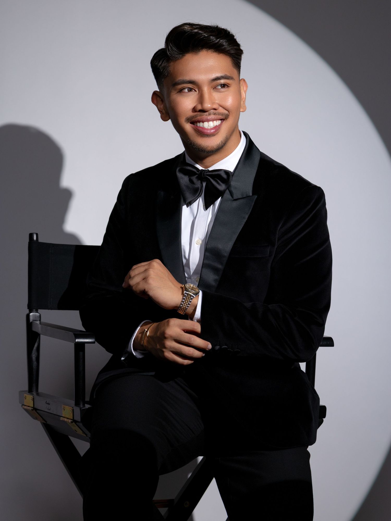 Professional studio portrait of person in black suit and tie seated in director's chair against gray backdrop with dramatic lighting.