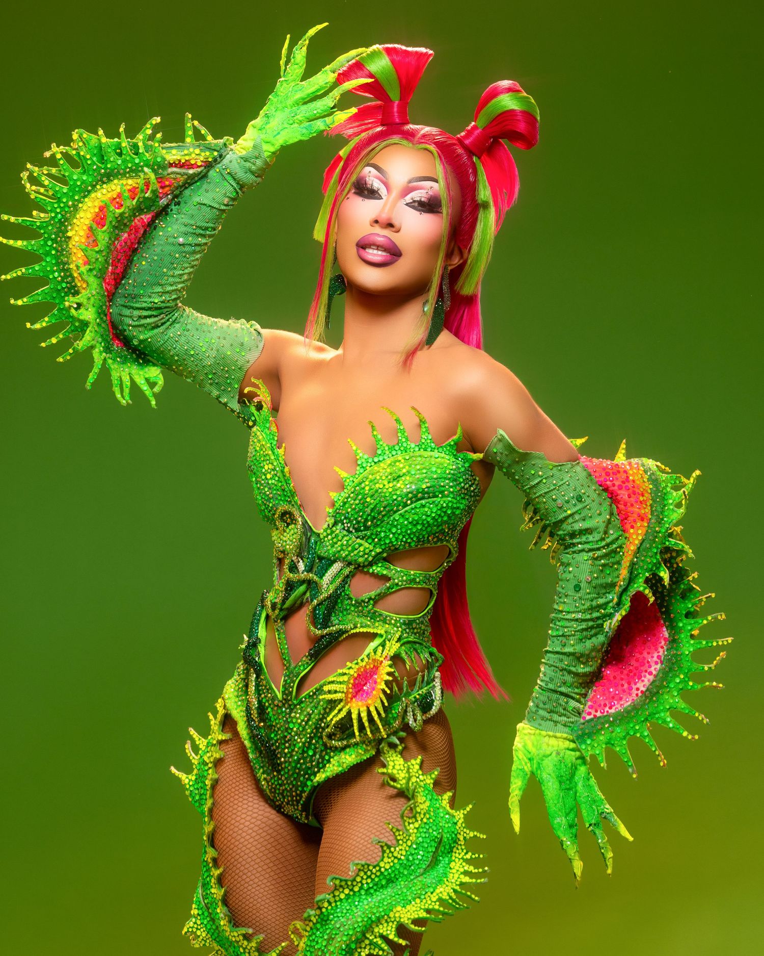 Performer in vibrant green Venus flytrap costume with pink hair and bedazzled bodysuit on green background.