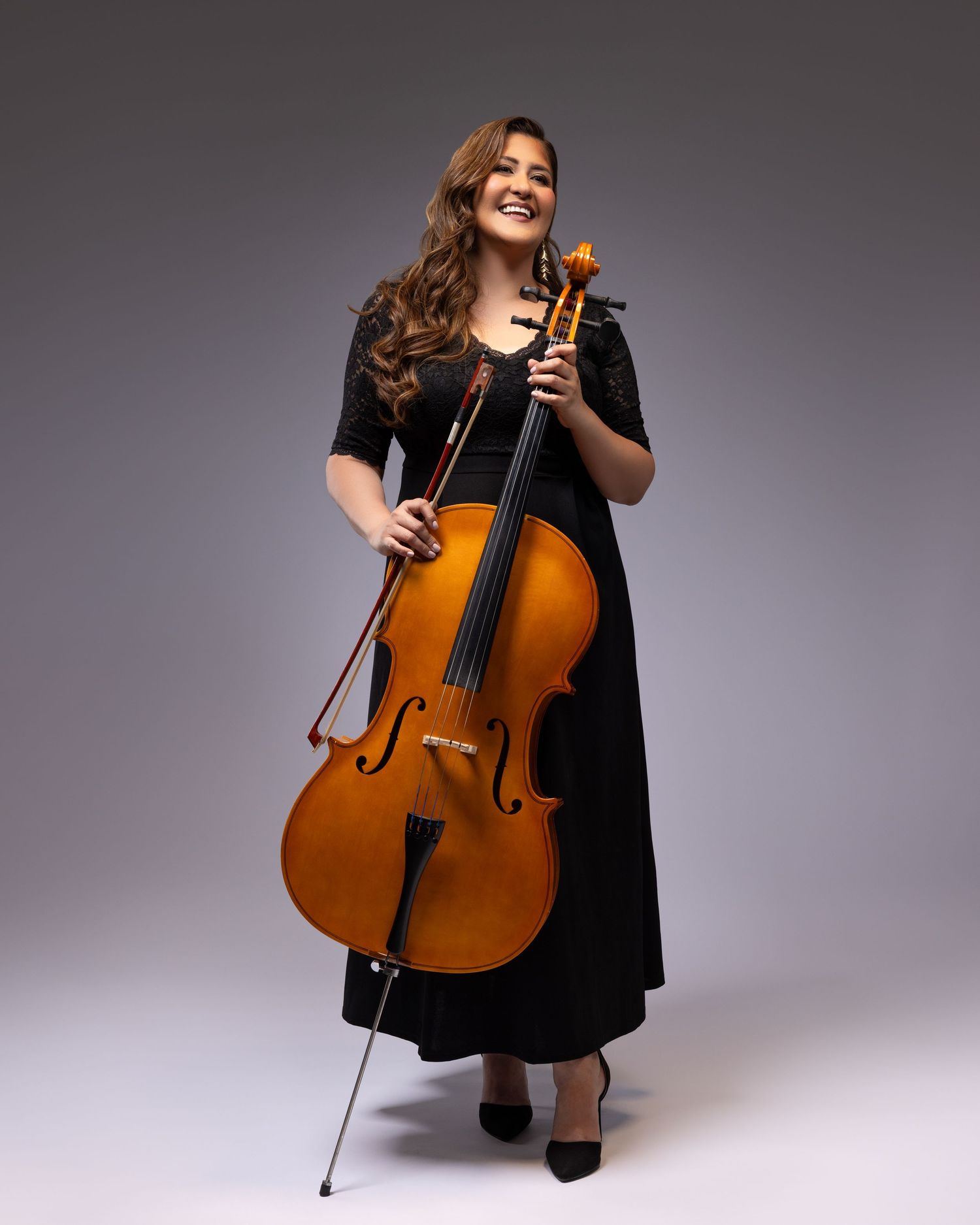 Musician in long black dress smiles while standing and holding a cello against gray studio background.