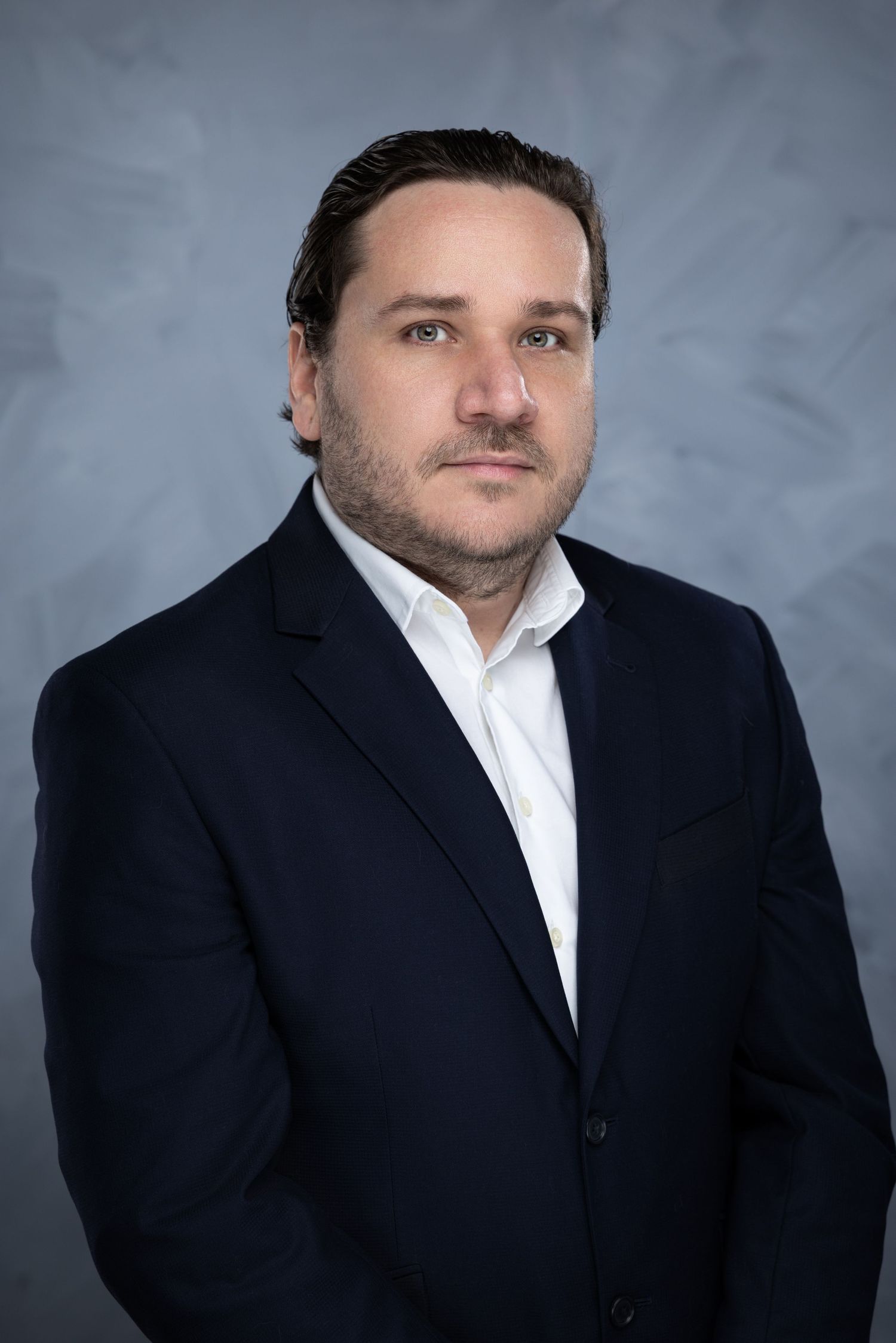Professional headshot portrait of person in dark suit jacket and white button-down shirt against gray background.