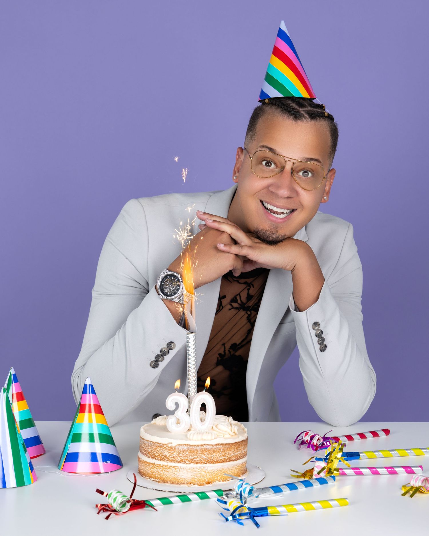 A person wearing a white blazer and party hat poses with a birthday cake and colorful party decorations on a purple backdrop.