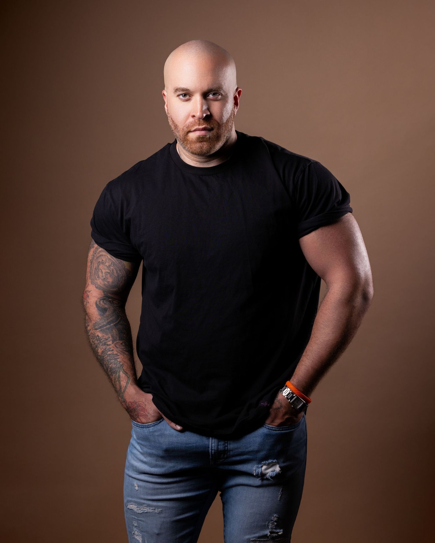 Man in black t-shirt and jeans poses against brown backdrop showing tattoos and confident stance.