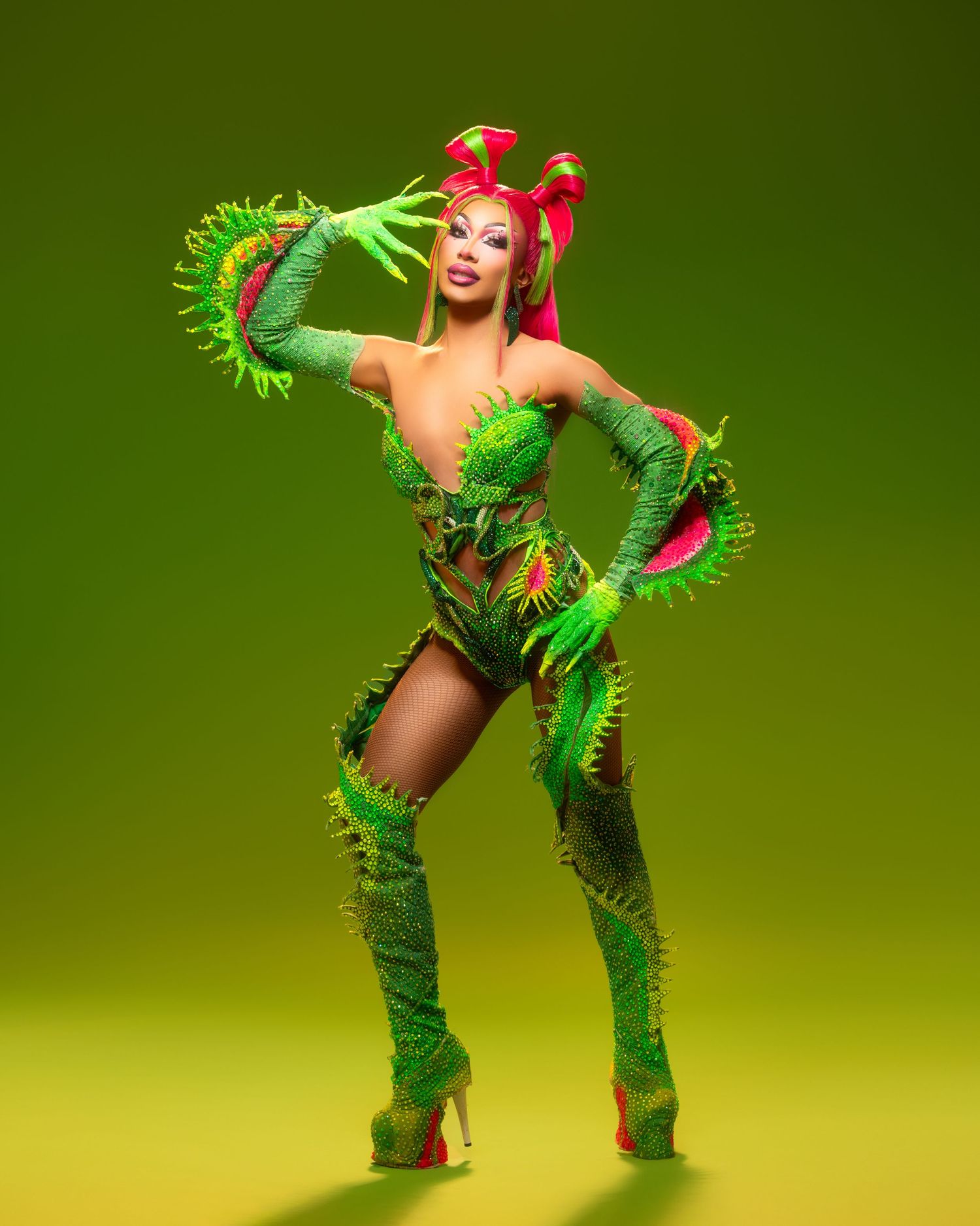 A performer in a bright green plant-inspired costume with feathers and boots poses dramatically against a green background.