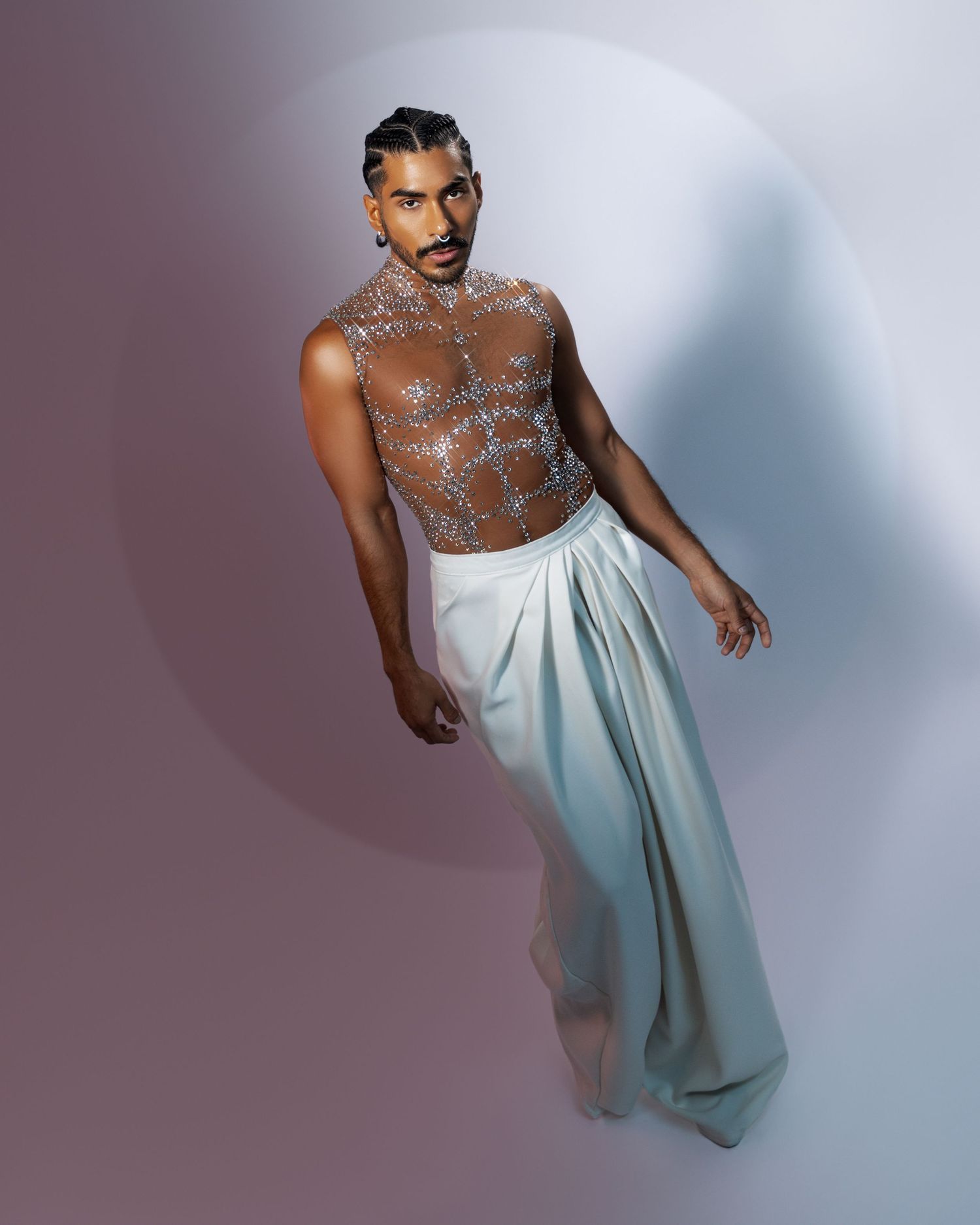 A performer in white flowing pants and glittery body paint poses dramatically against a gradient background.